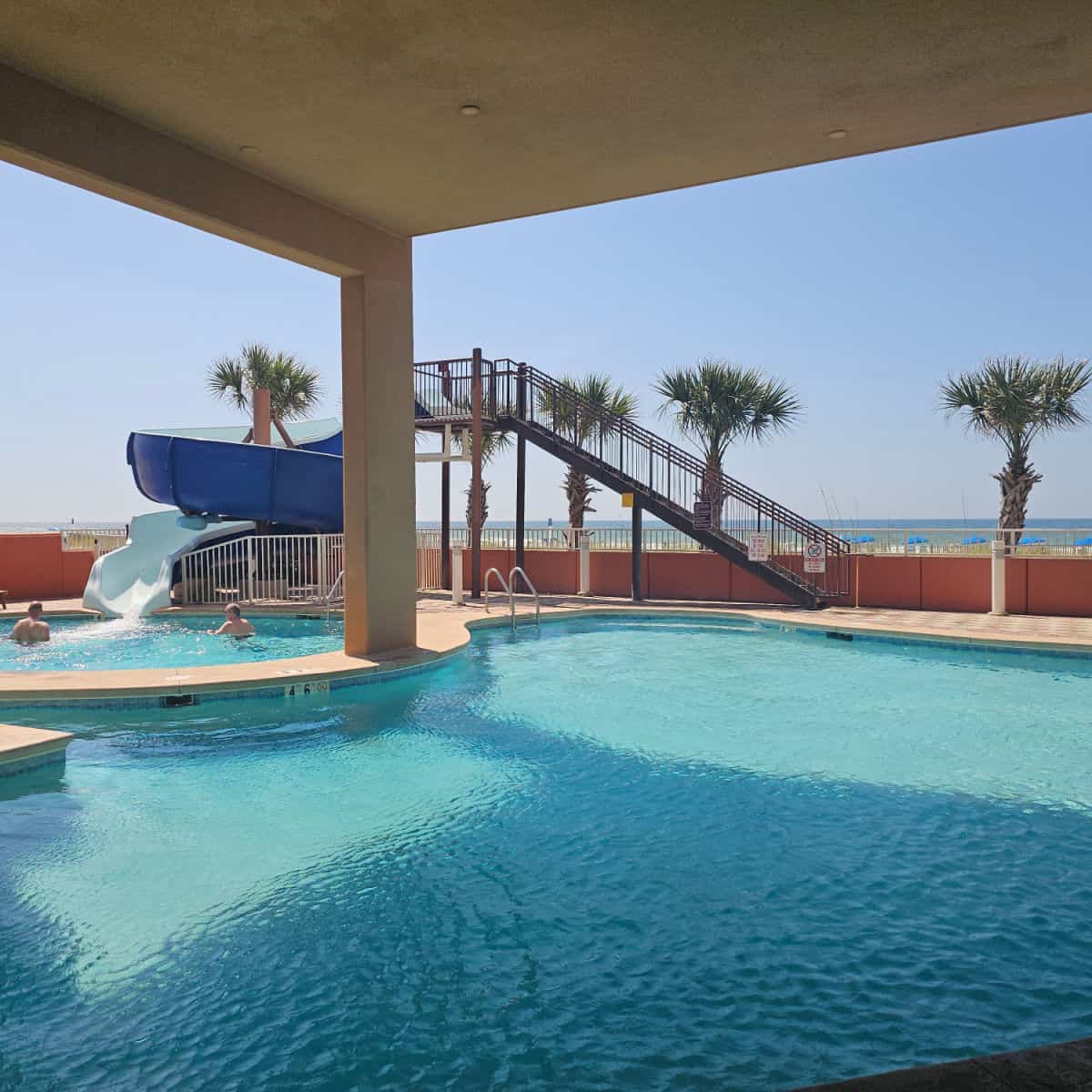 pool with a waterslide in the left side with two people in the water looking out towards the Gulf