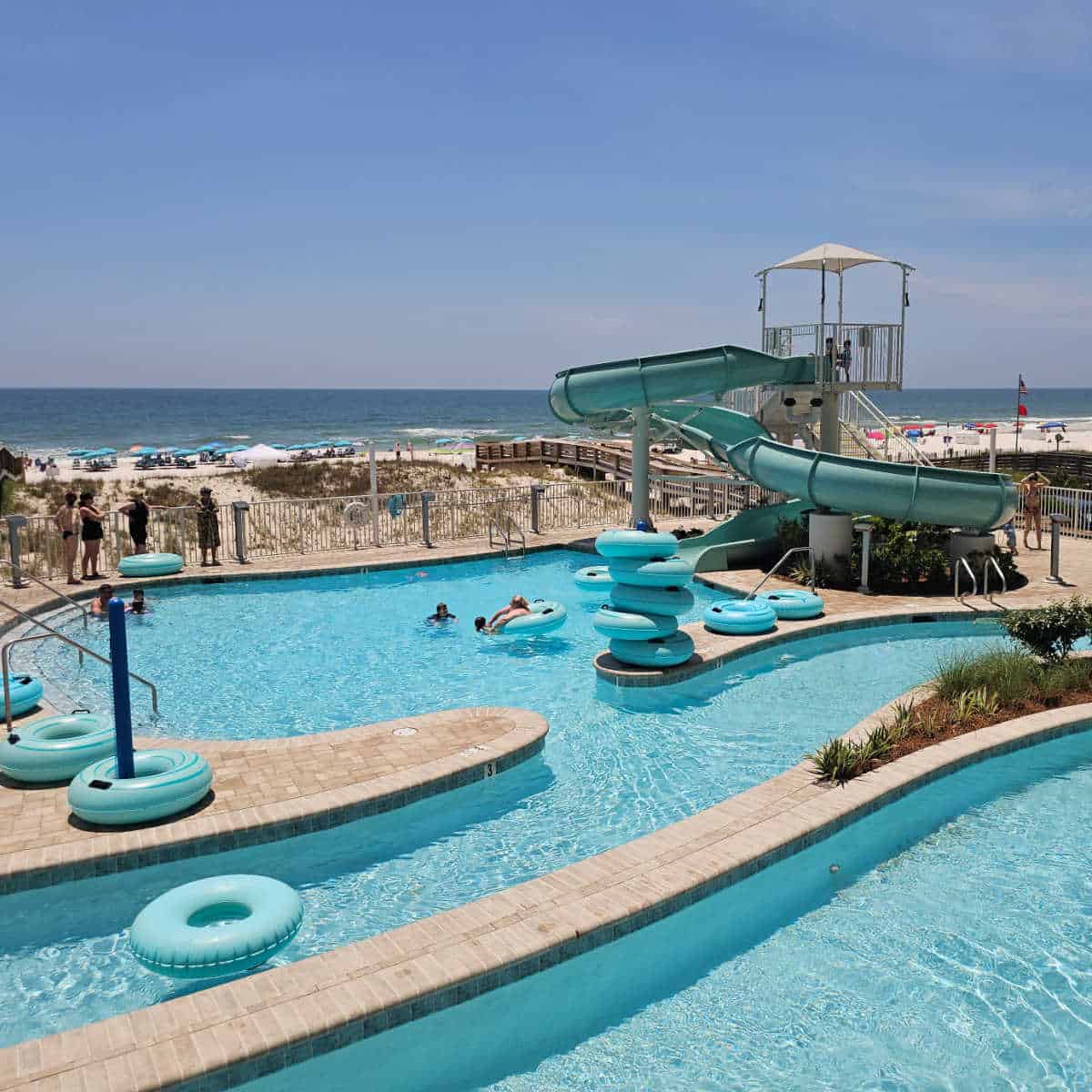 waterslide leading down to a pool with a lazy river, intertubes floating on the water and set up to the side, looking towards the beach with sand dunes and Gulf water