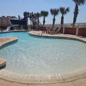 zero entry pool with a waterslide in the background, deck chairs on the pool deck, palm trees a bit of gulf waters in the background