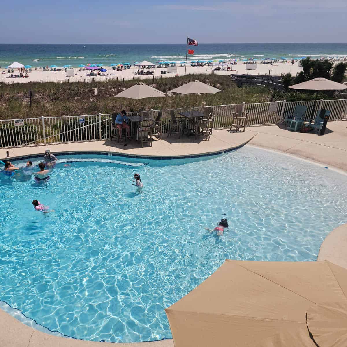 Zero entry pool with kids playing in the water, covered table and chairs along the pools edge, looking out toward the beach with white sand and Gulf waters