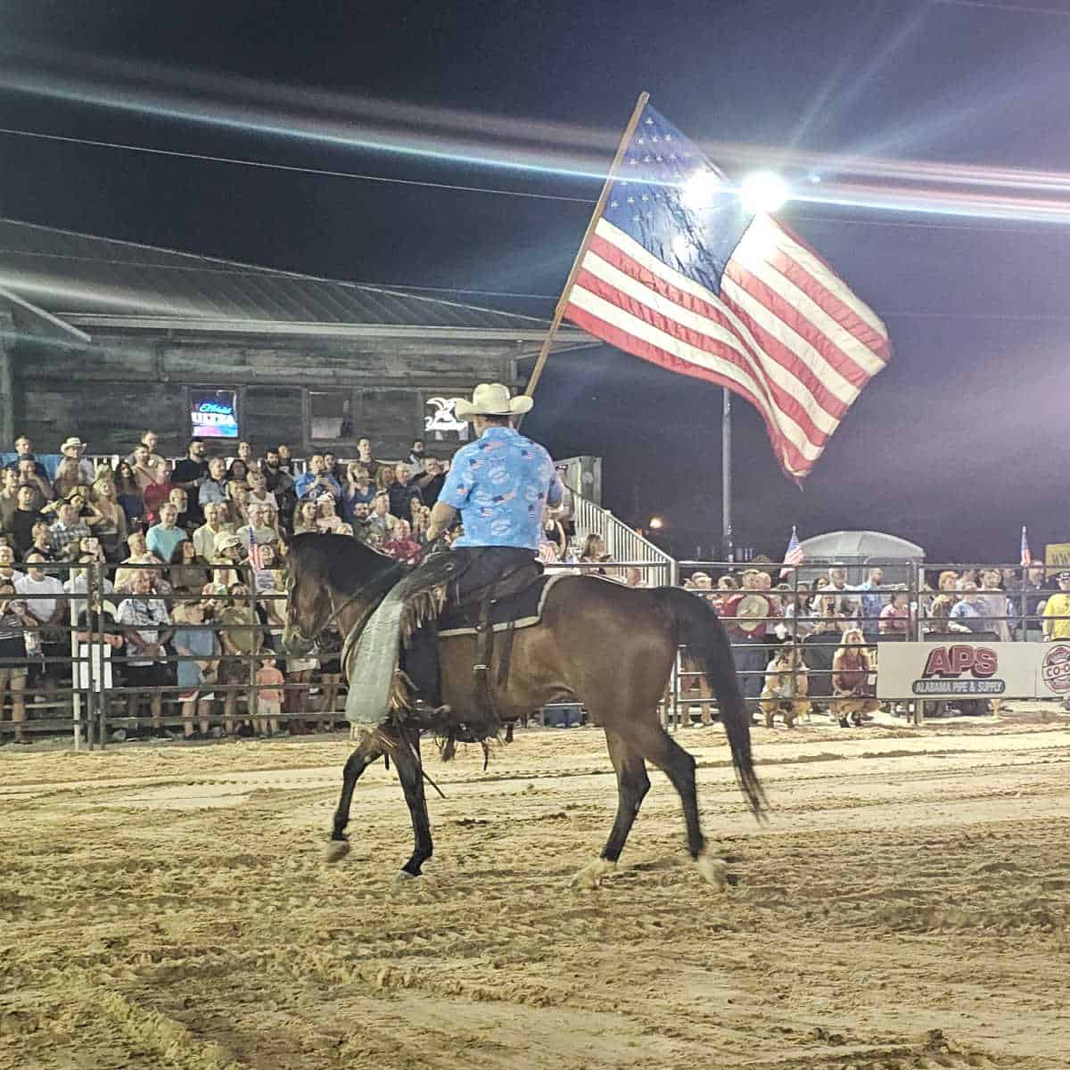 cowboy on a brown horse carrying an American Flag with people standing in bleachers and the Flora Bama in the background