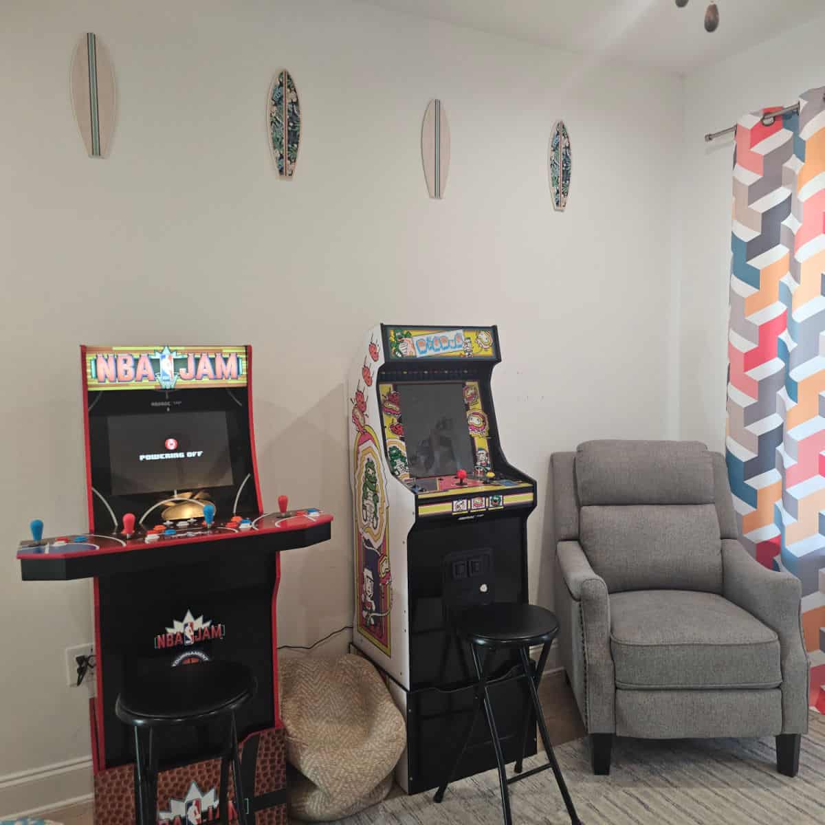 Arcade games with stools next to a chair, surfboard art on the wall