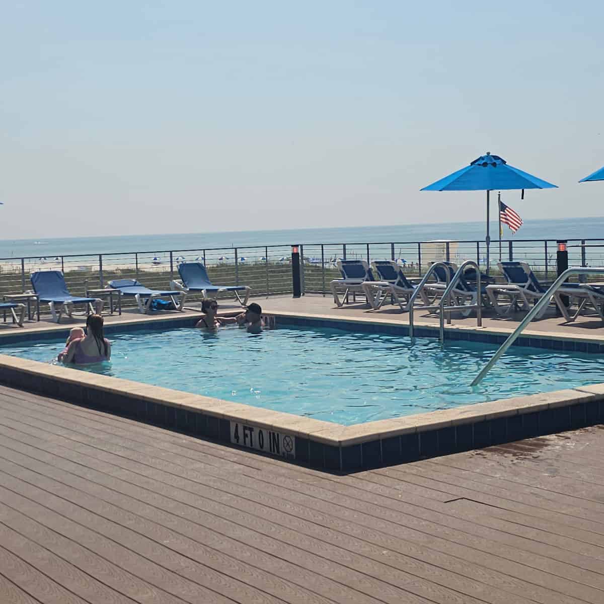 outdoor pool on a deck next to lounge chairs and a blue umbrella looking out to the Gulf