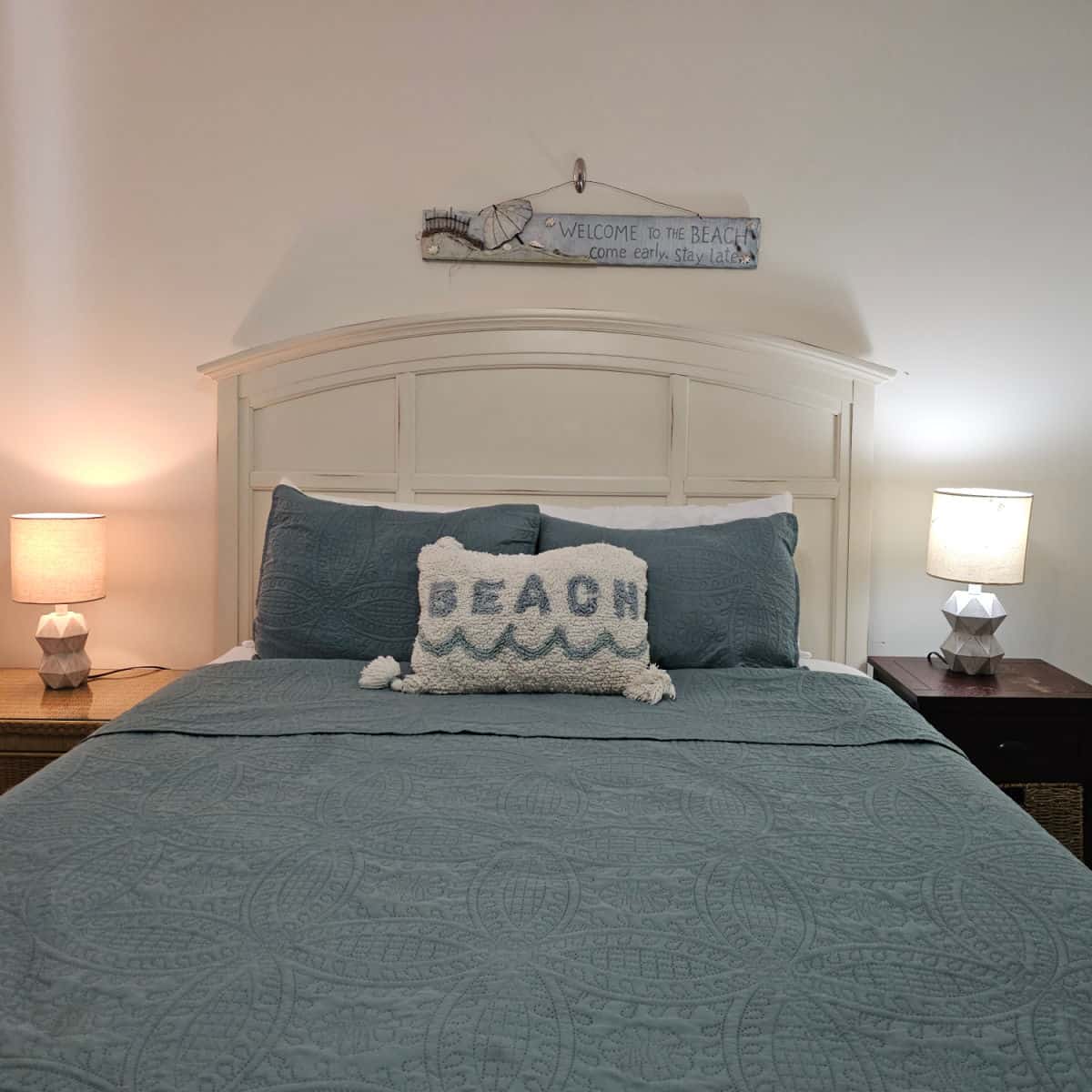 Bedroom with bed with beach pillow, two nightstands with lamps, welcome to the beach sign over the bed