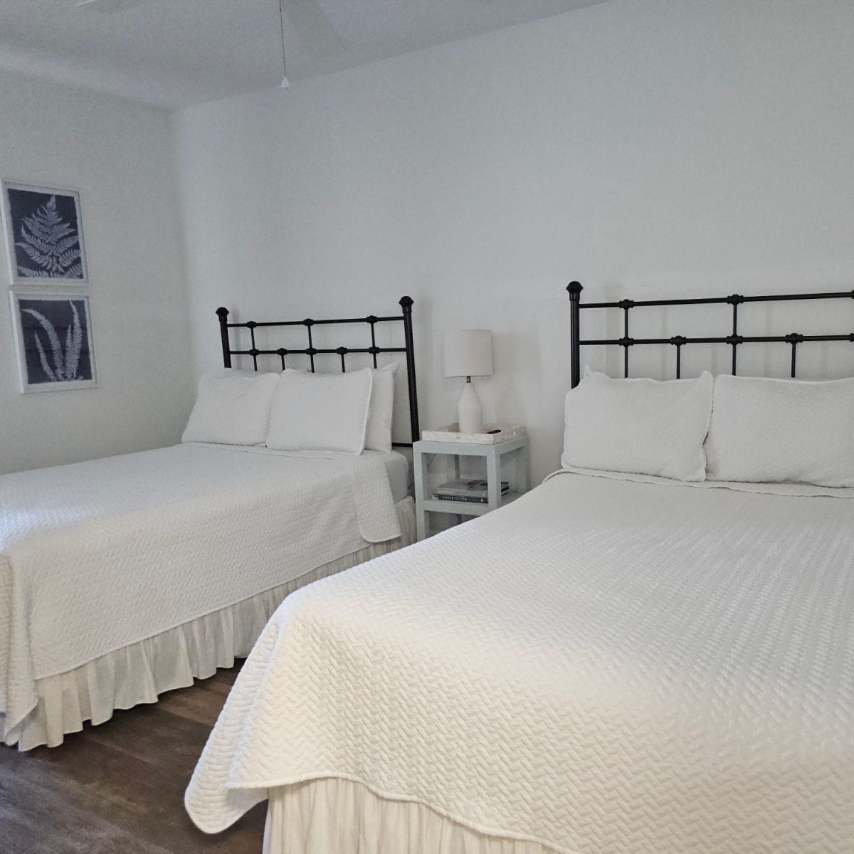 two beds with metal frames, table in between the beds with a lamp on it. white walls with two leaf prints