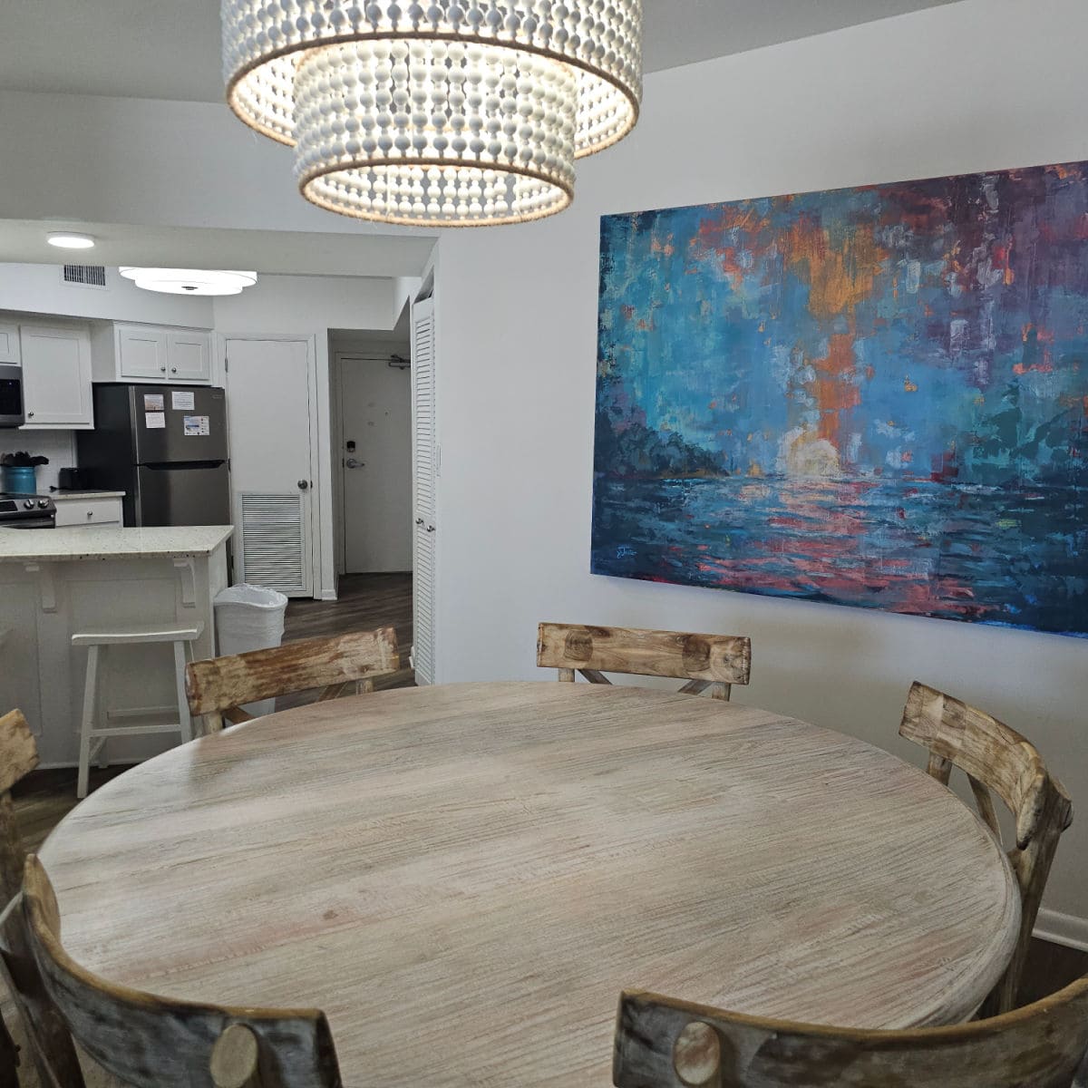 large circular wooden dining room table with chairs around it, colorful painting on the wall, looking towards the kitchen on the far left side.