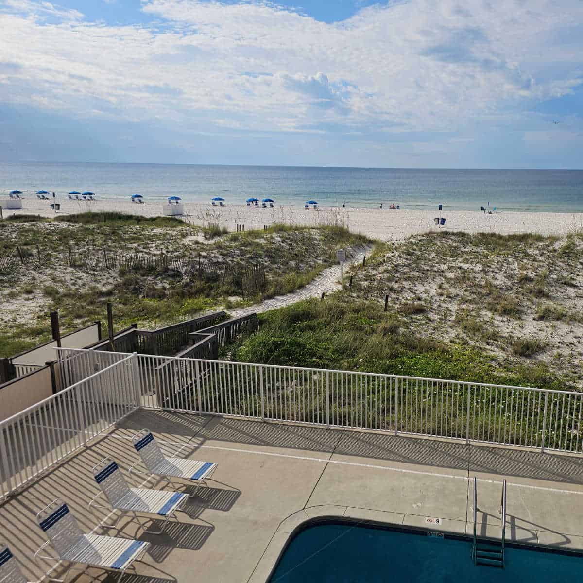 edge of an outdoor pool and pool deck with lounge chairs surrounded by a fence, sand dunes and a sand walkway leading to the beach and Gulf waters