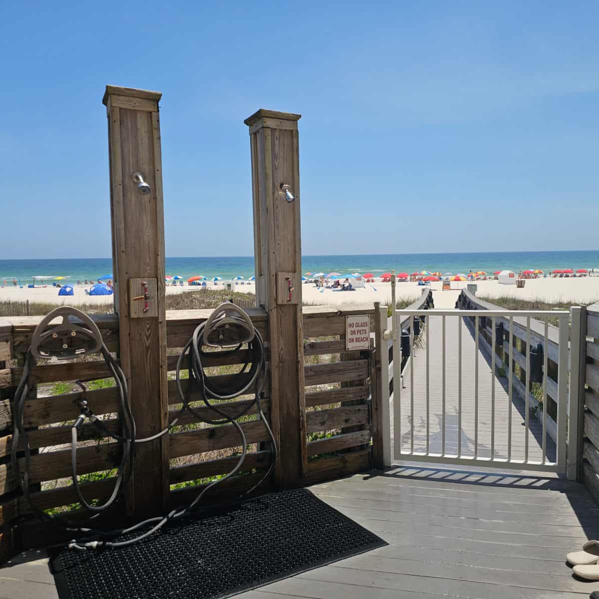 two outdoor showers and hoses on a wooden fence with a gate leading out to the beach over a boardwalk, Gulf water in the background