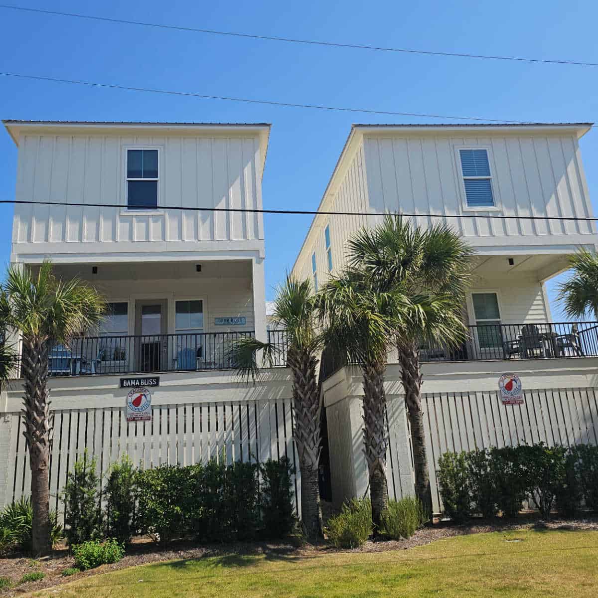 Bama Bliss sign and Beachball Properties signs on the side of two story vacation rentals with palm trees in front of them