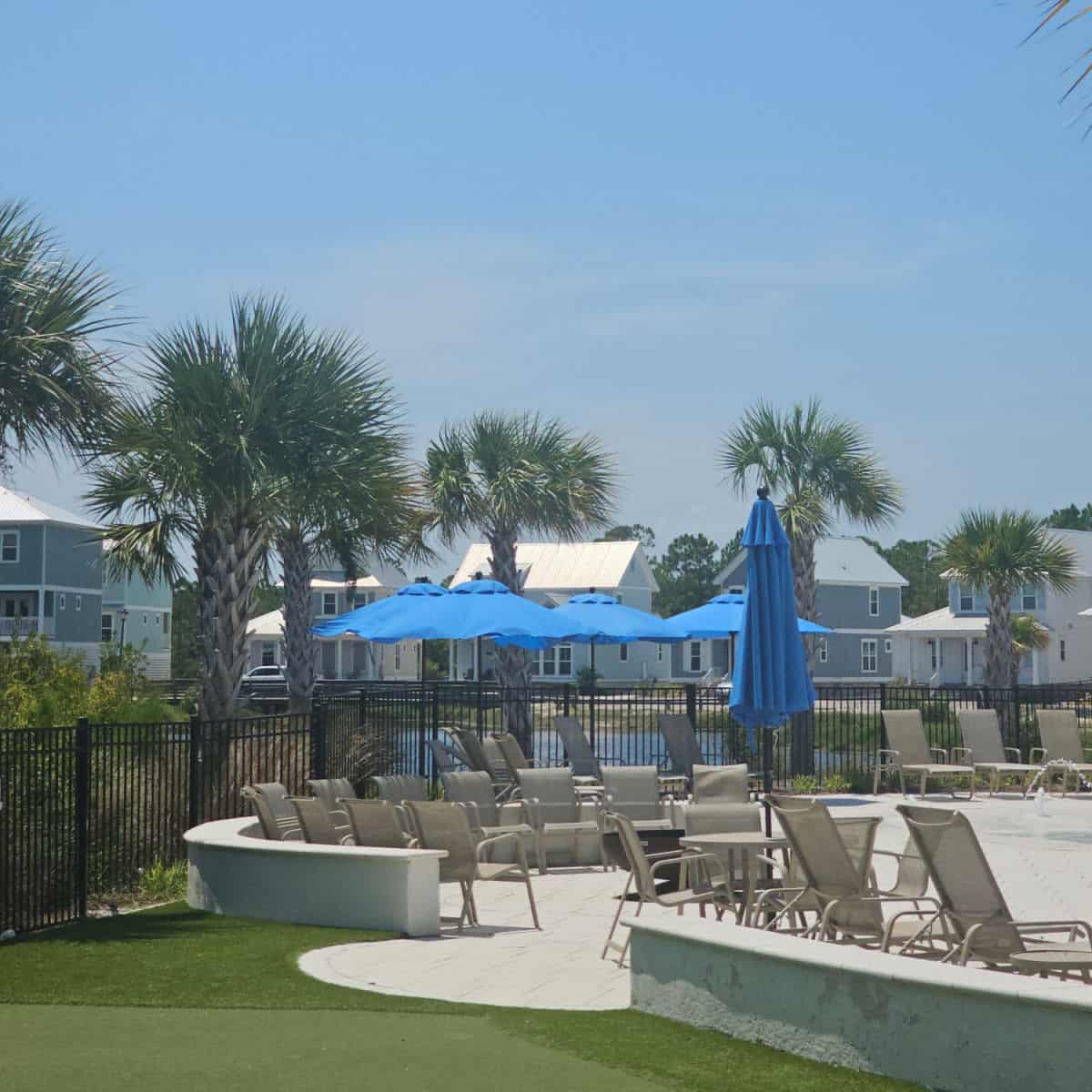 lounge chairs on the pool deck with blue umbrellas, palm trees by the fence