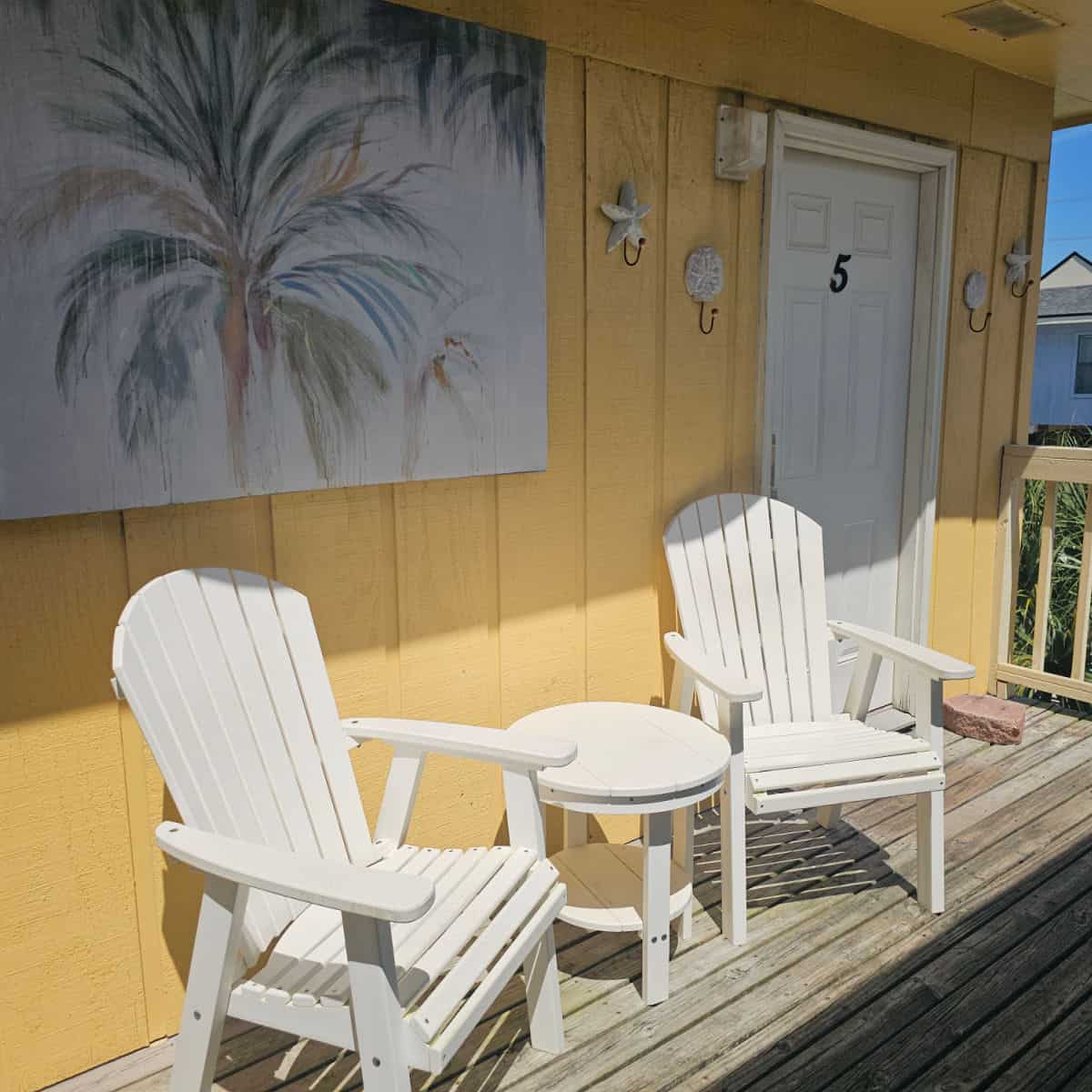 yellow house with a white door with 5 on it, 2 white Adirondack chairs and table on the deck, palm tree print on the side of the house