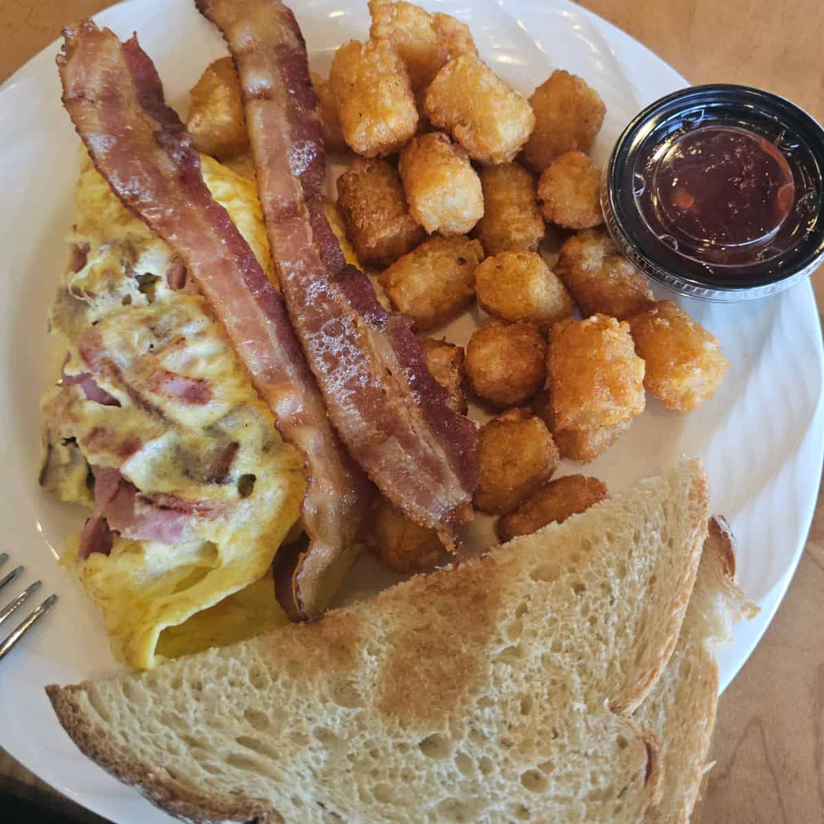 breakfast plate with omelet, bacon, tater tots, toast, and a container of salsa on a white plate