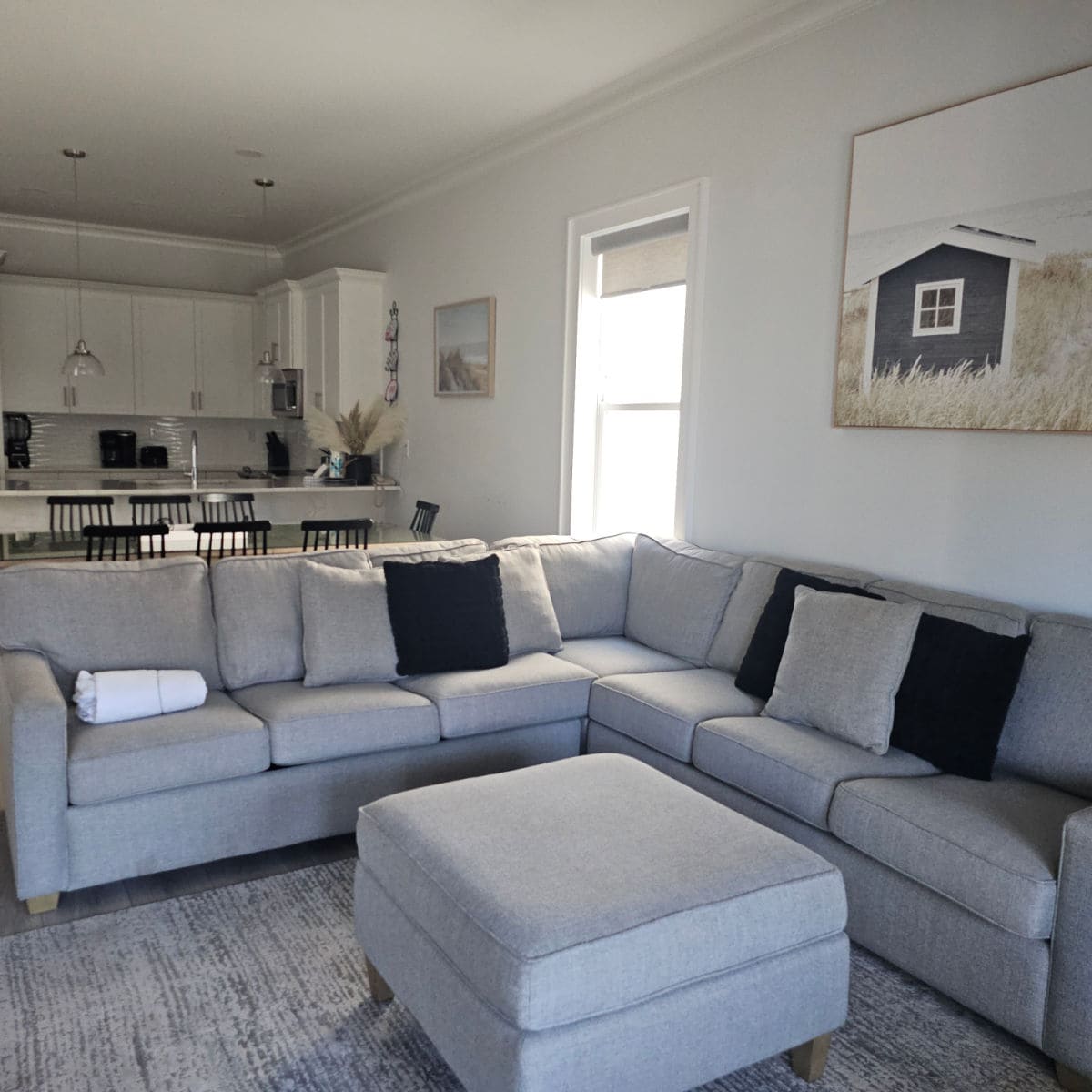 Living room with wrap around couch with lots of pillows and an ottomon. Behind the couch is the kitchen with white cabinets and pendant ligths