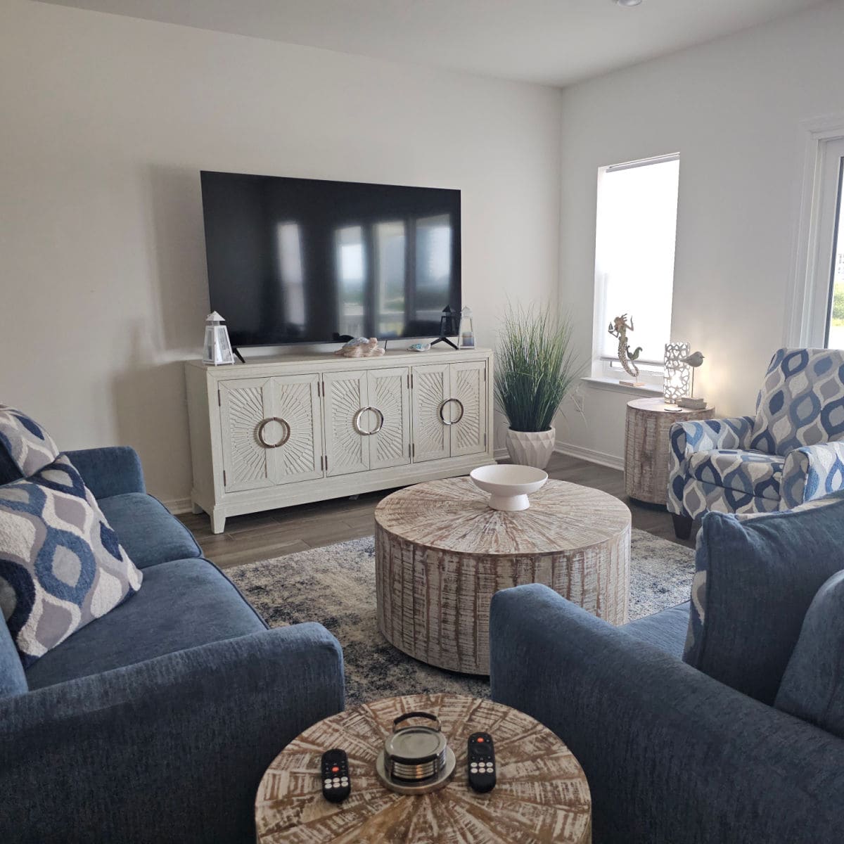Living room area with chairs and couch around a circle wood table with a white bowl on it, tv on a cabinet near the wall