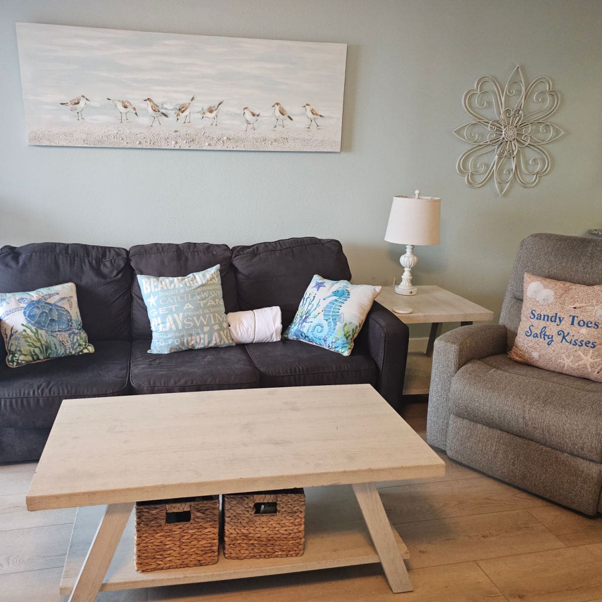 Living room with a brown couch, ta chair, and colorful coastal pillows, shore bird painting on the wall with a table and two baskets on the floor