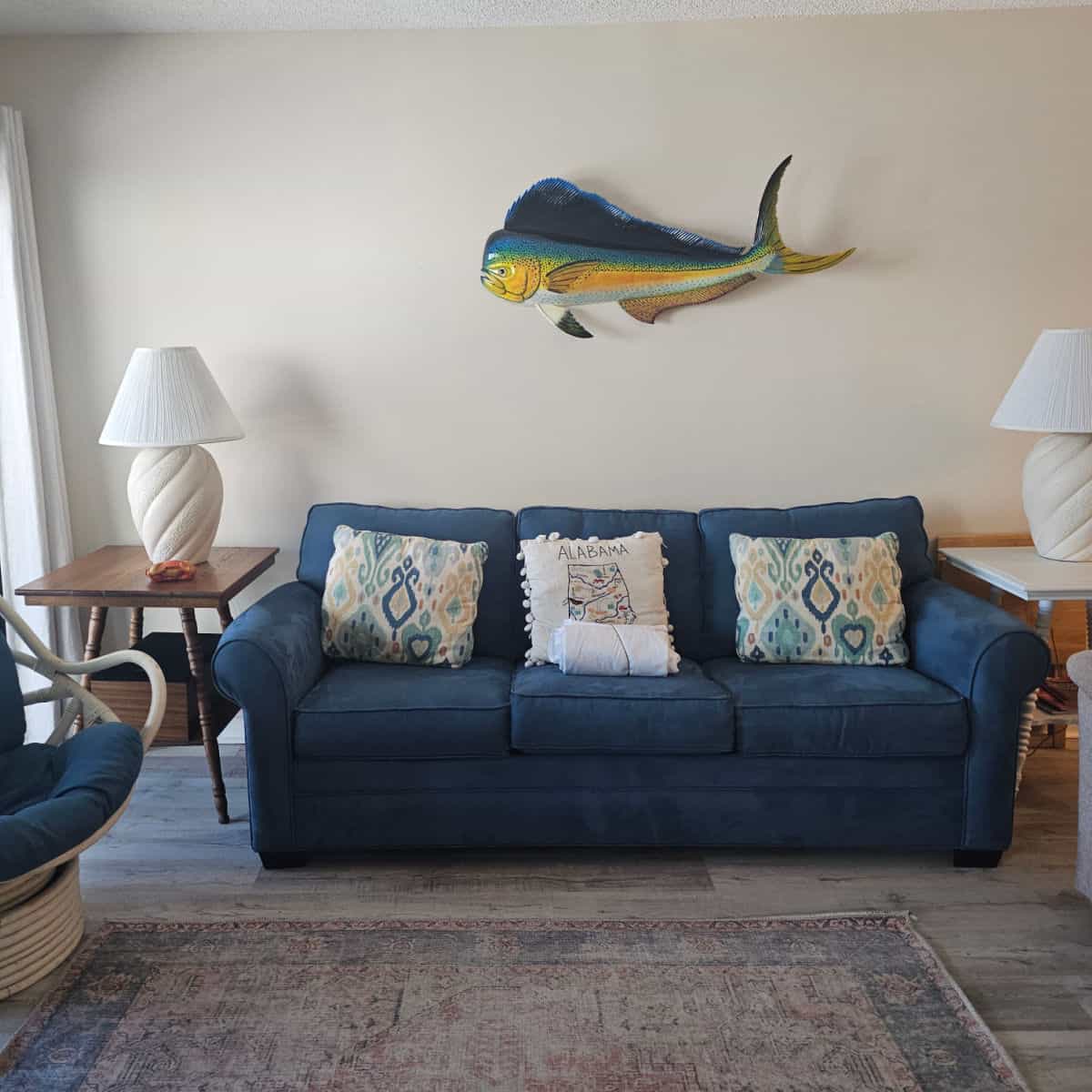 Blue couch with an Alabama pillow and two colorful pillows, large fish above the couch, tables on either side of the couch with lamps