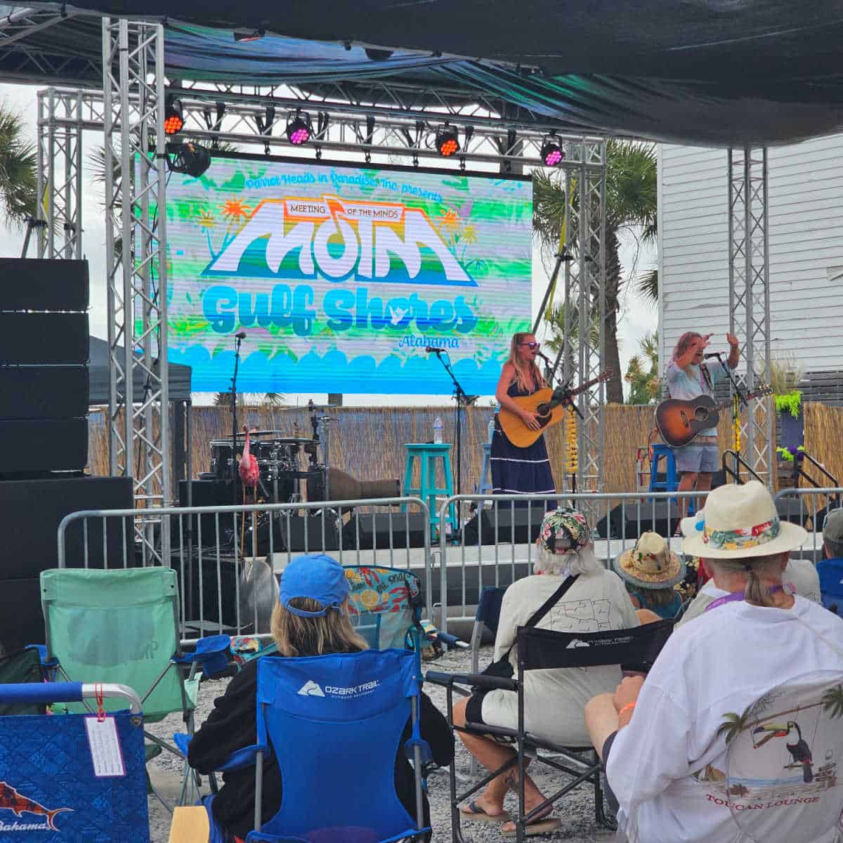 Meeting of the Minds Gulf Shore Alabama sign behind a female singer on stage and a guy with a guitar, people sitting in camp chairs watching