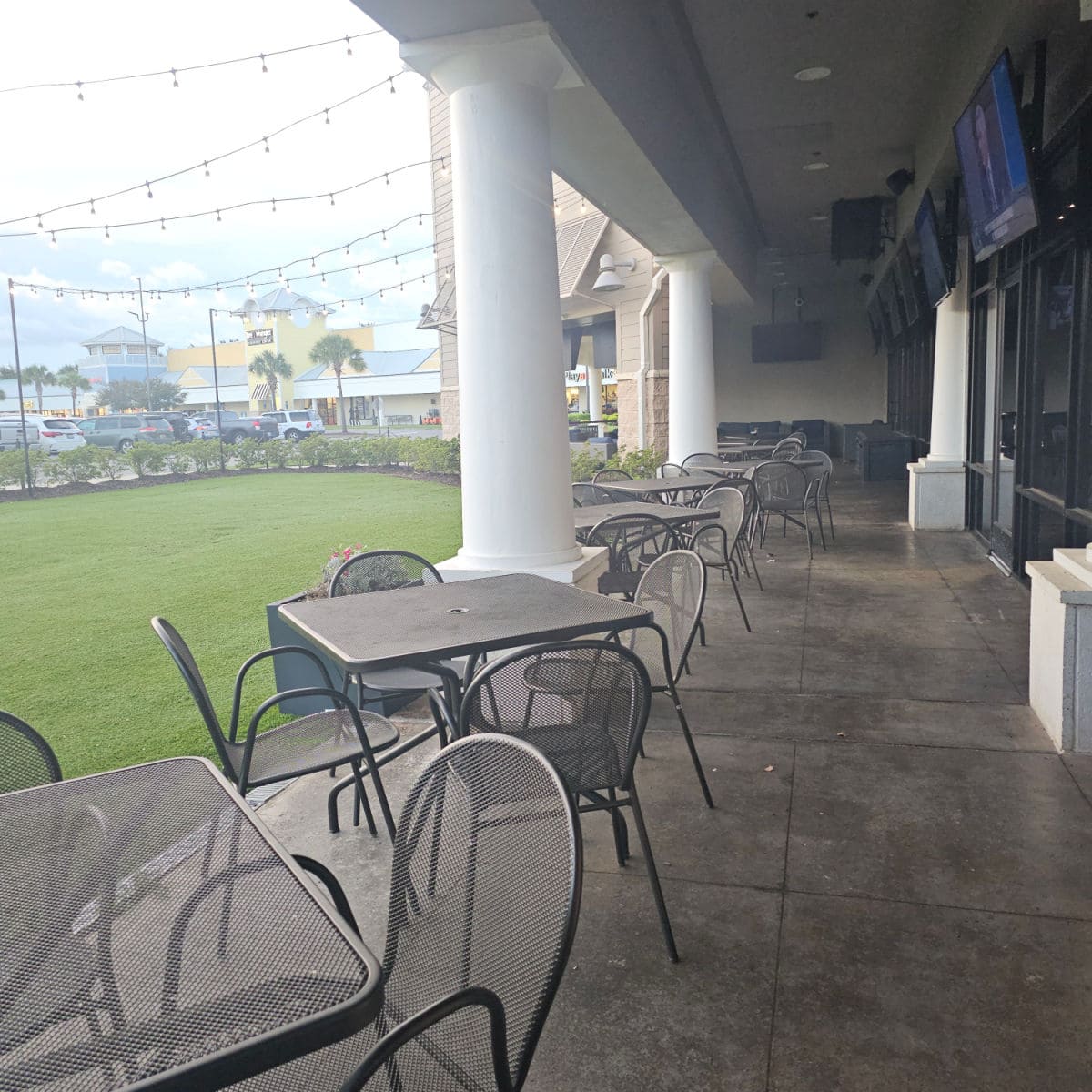 outdoor tables and chairs under a cover with TVs mounted on the wall next to the restaurant, green lawn with lights above it to the left and views of Tanger Outlet stores
