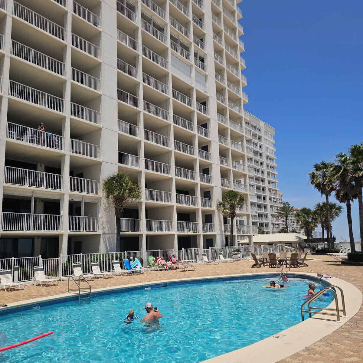 Outdoor pool with people in the water, lounge chairs around a pool deck, tall Windward Pointe building next to the pool