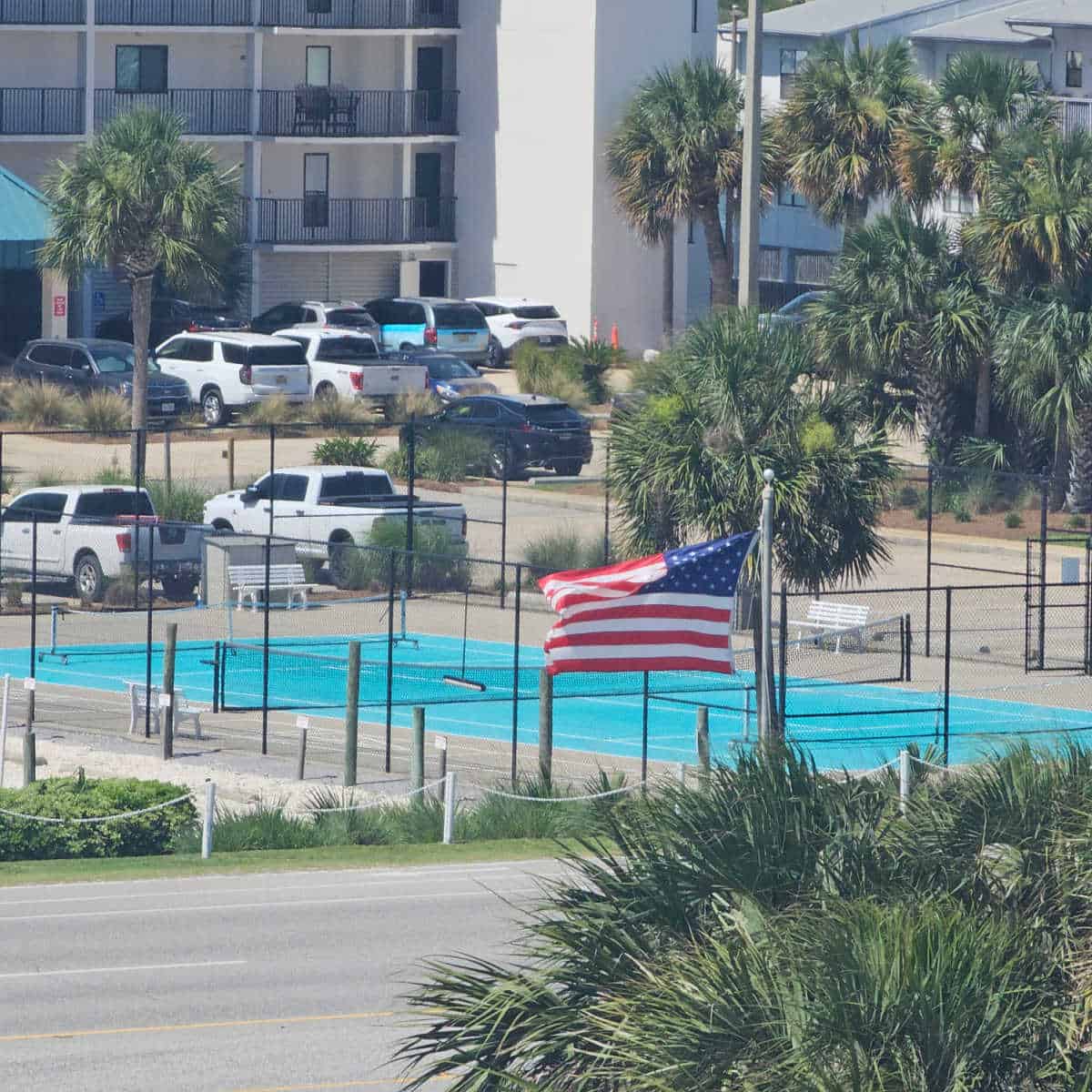 Tennis court behind a fence with cars parked in the parking area next to Wind Drift condo buildings