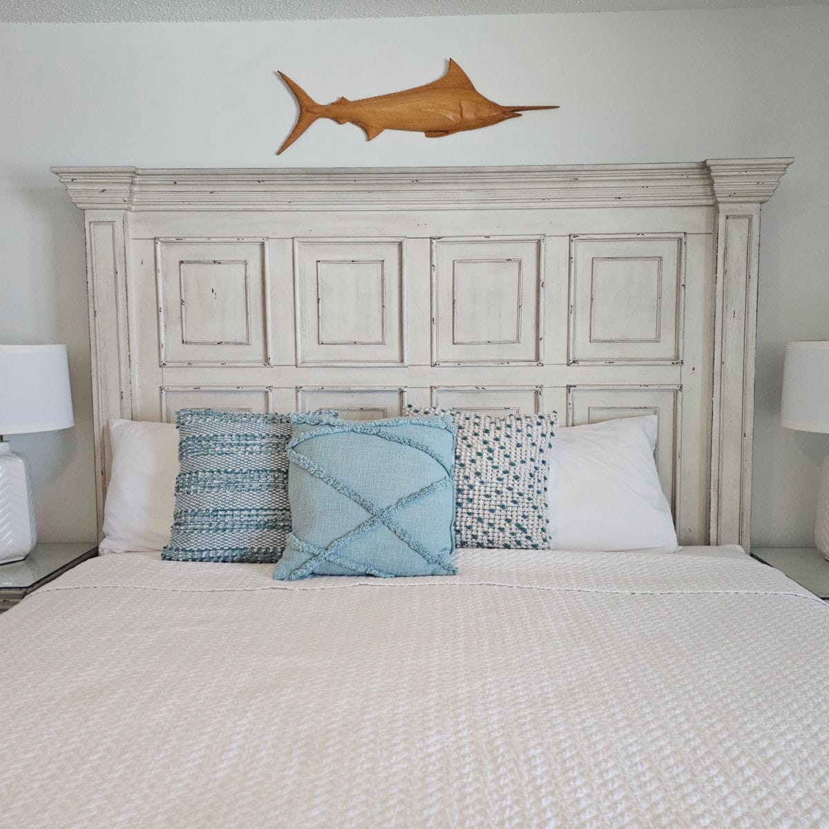 Bed with wooden bed frame, wooden marlin fish over the bed, white lamp and shade to the left of the bed