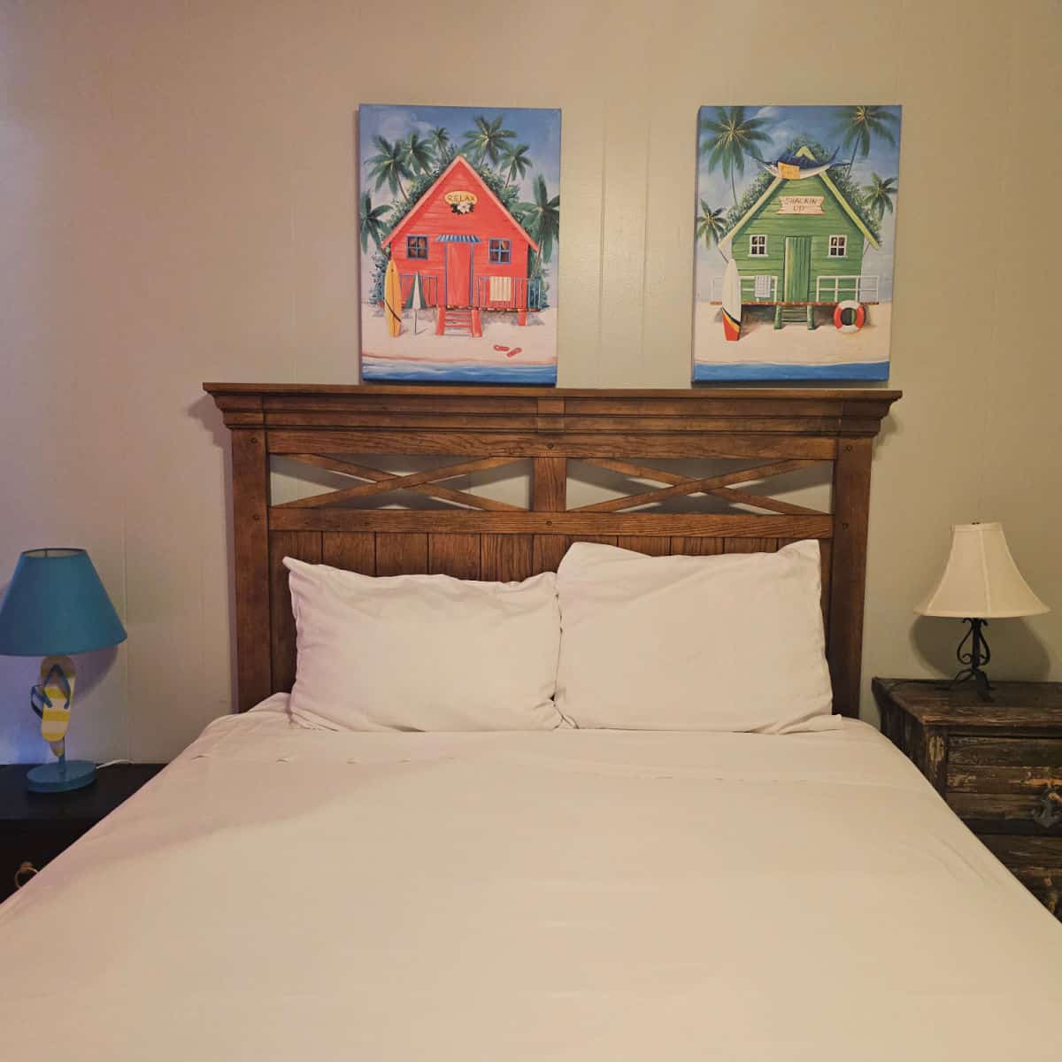 Bedroom with wooden frame and white linen, nightstand with lamps, two tropical prints over the bed.