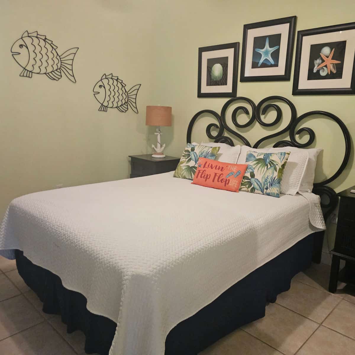 bedroom with white linen covered bed with tropical pillows, shell art over the bed and metal fish art on the far wall