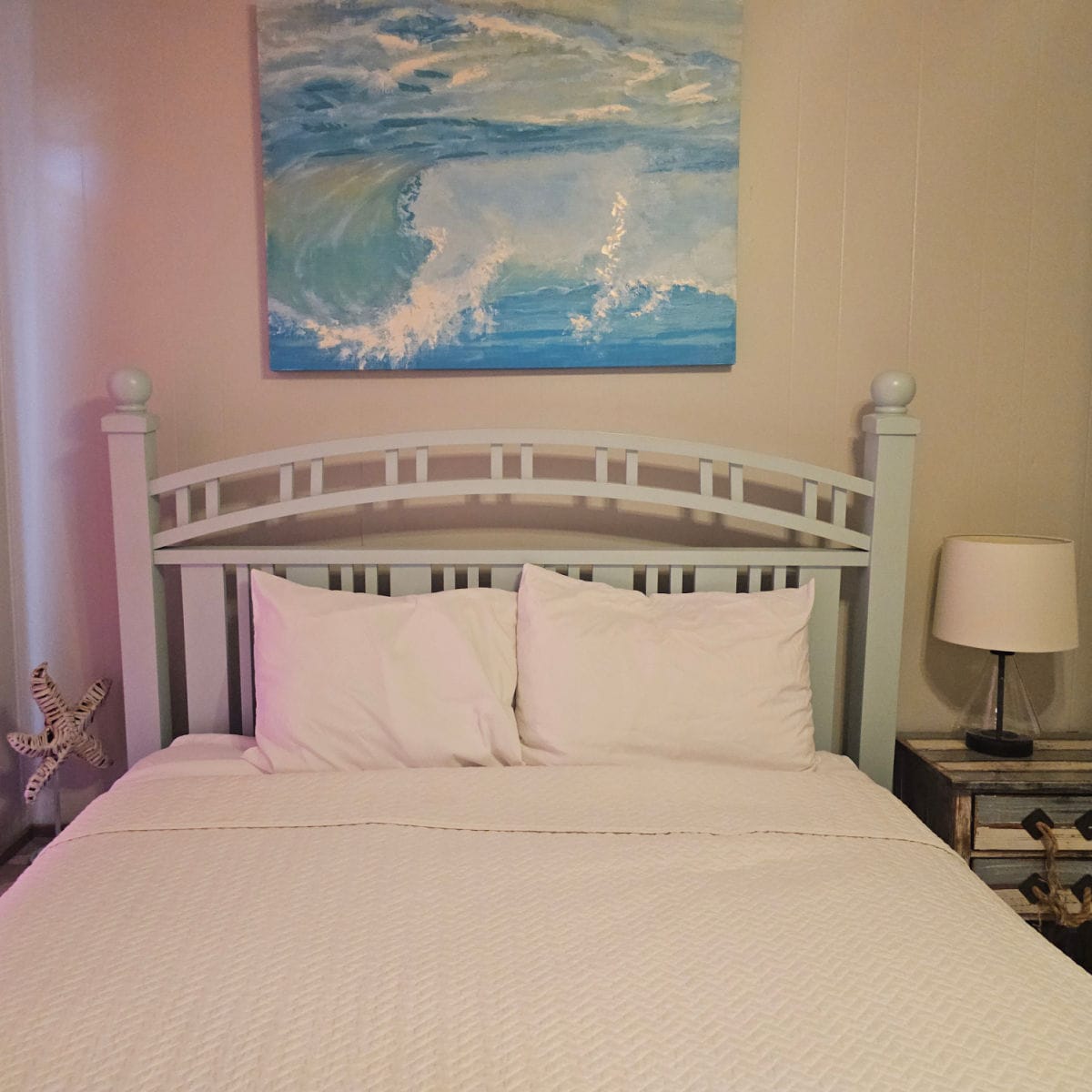Bedroom with a wooden bedframe, white linens, nightstand with lamp, and blue wave photo over the bed