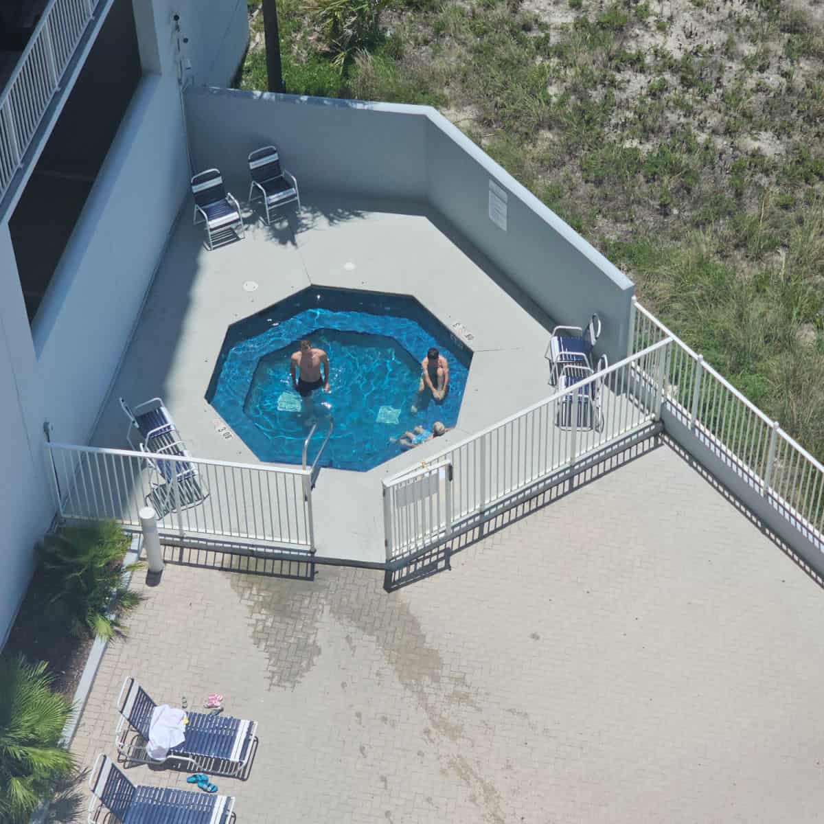 hot tub behind a fence with a couple of people sitting in it, chairs around the deck