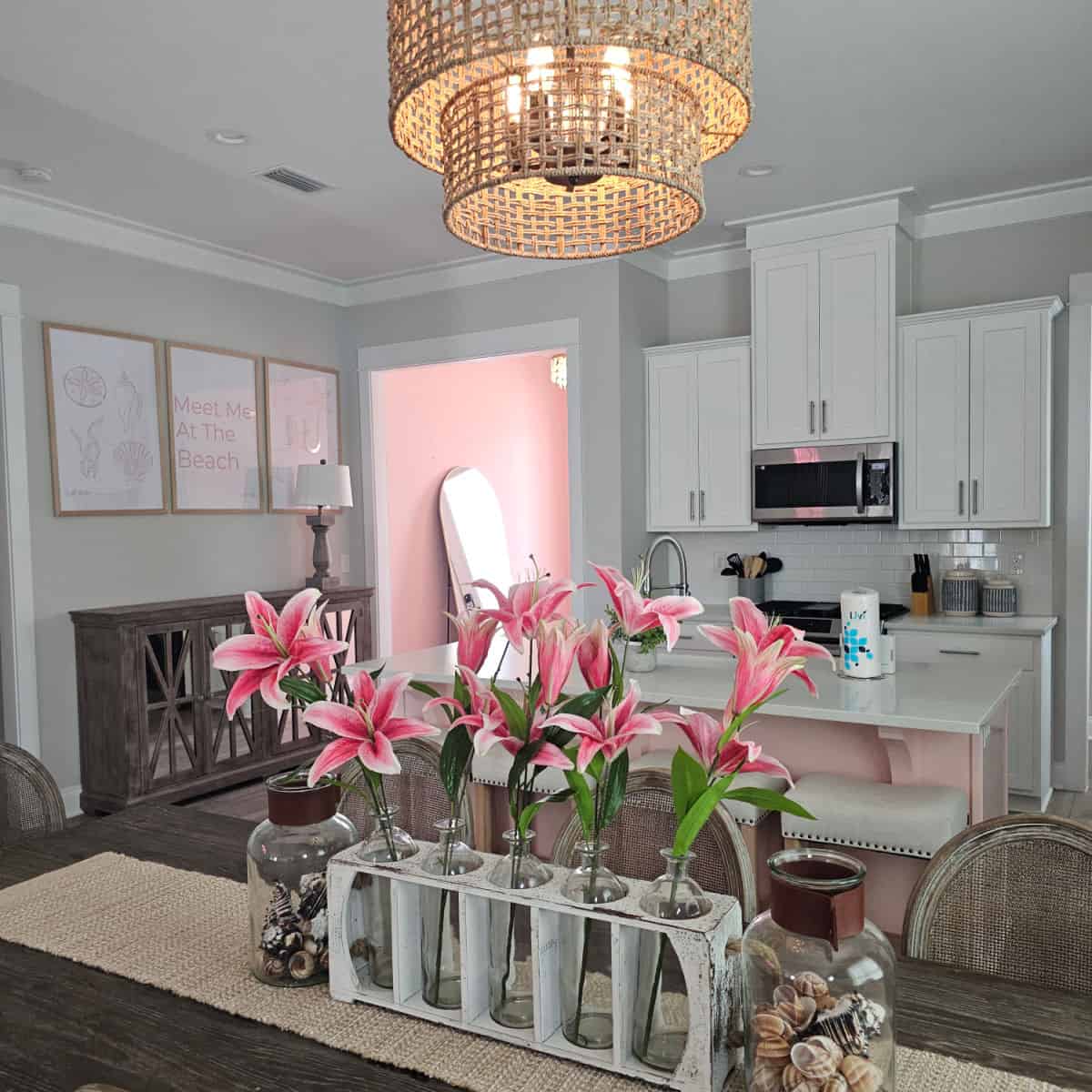 Pink lily in vases on the dining room table looking over to the kitchen with white cabinets and stainless steel appliances. Meet me at the beach art on the wall near the pink entrance way with a mirror