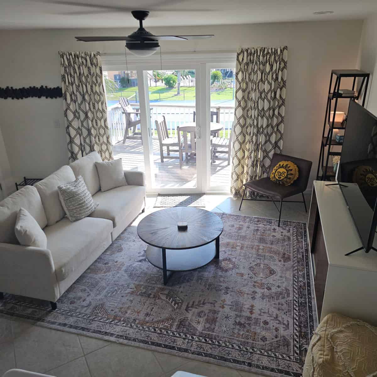 Looking down into the living room with a cream colored couch, brown chair with sun pillow, wooden circle table, and out to a balcony with table and chairs