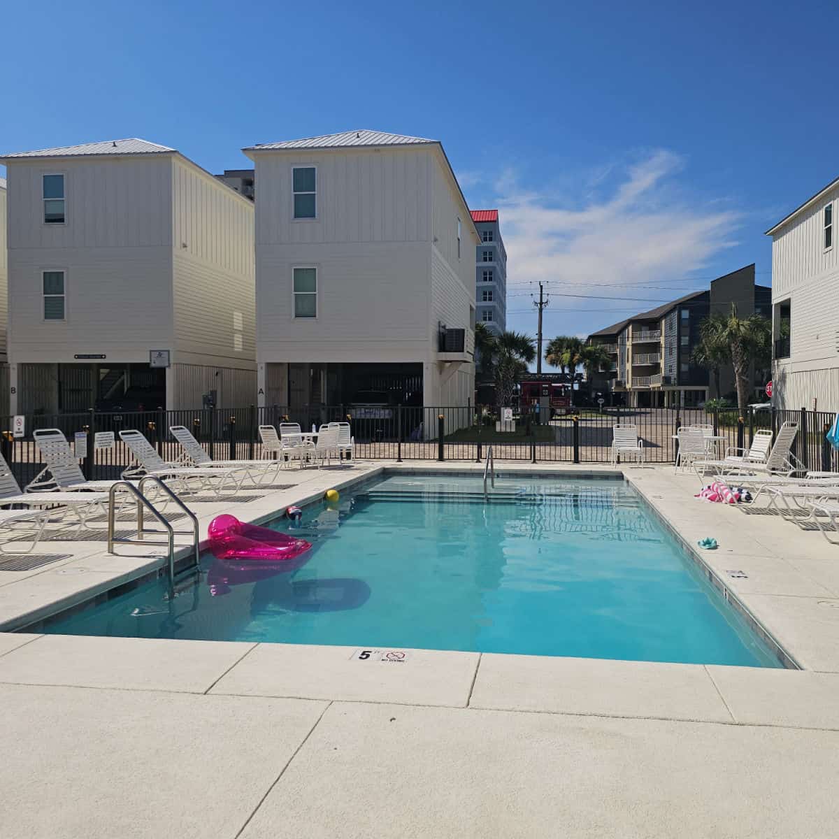 outdoor pool area with floaties in the water, lounge chairs around the fenced in pool deck. Pelican Place Cottages buildings around the pool
