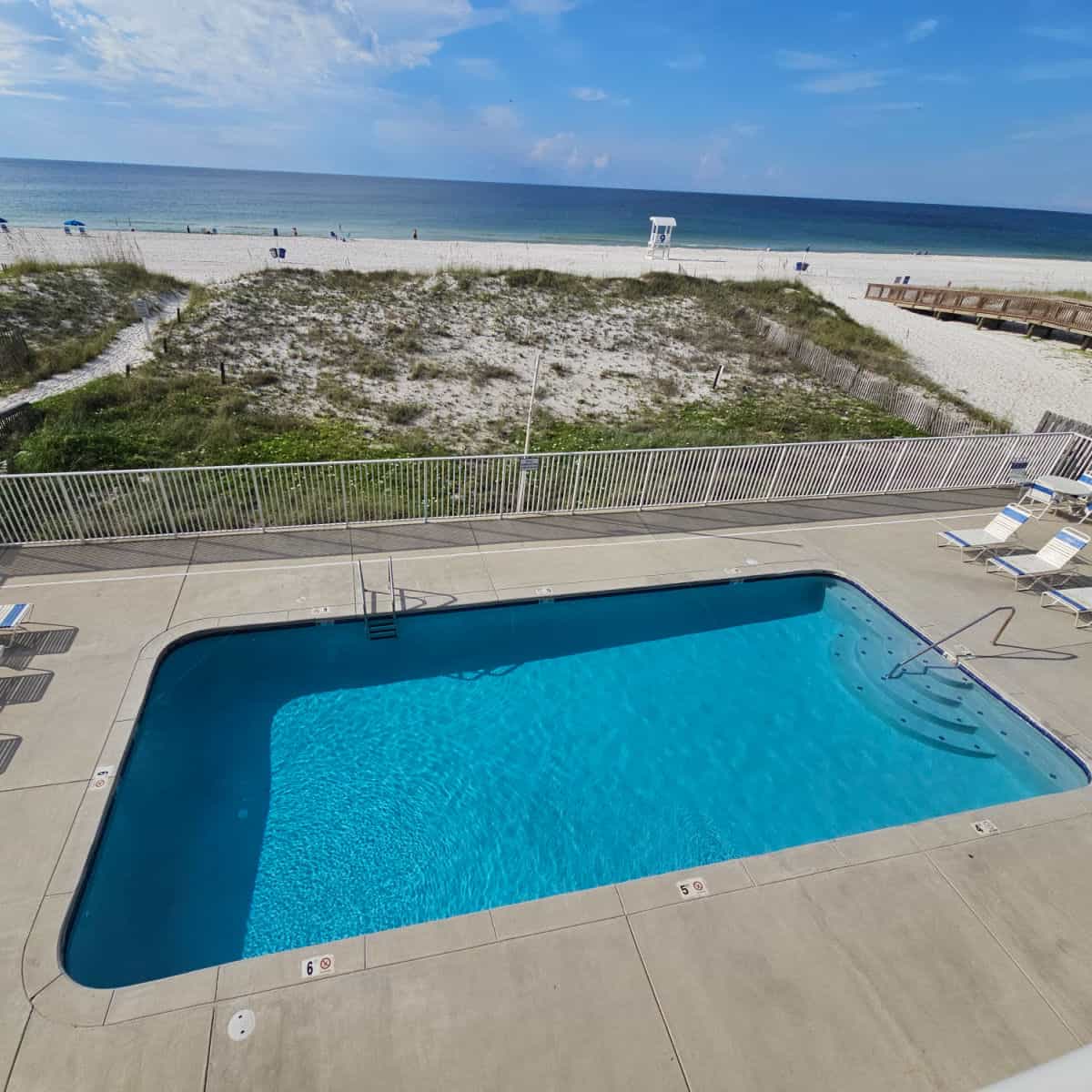 Outdoor pool with lounge chairs around it white fence leading to the sand dunes and beach with Gulf waters and a lifeguard stand