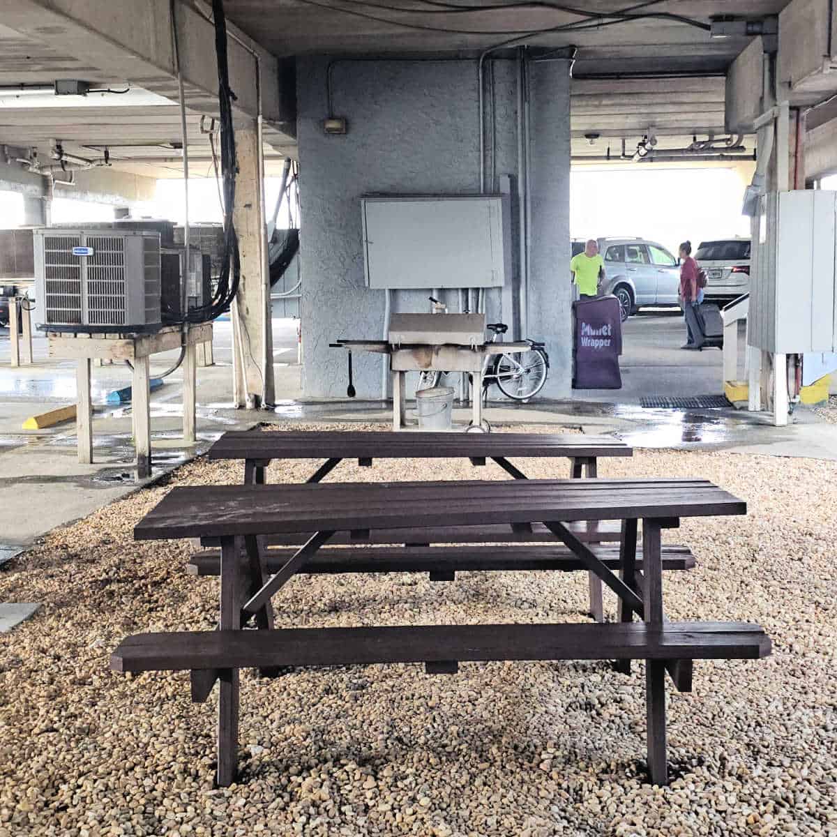two brown picnic tables on gravel with a charcoal bbq near a wall