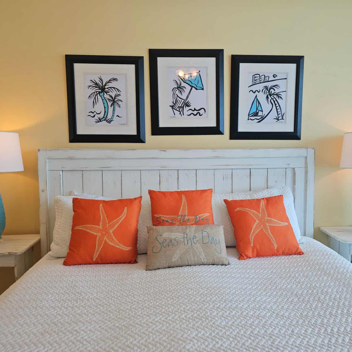 bedroom with white linens and orange pillows with star fish and a seas the day pillow, colorful art on the wall above the bed, white lamp shade in the corner.