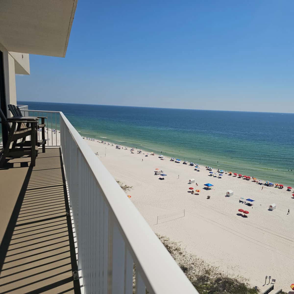 balcony with two tall adirondack chairs over looking Orange Beach beach with umbrellas spread across the sand and the Gulf on a clear sunny day