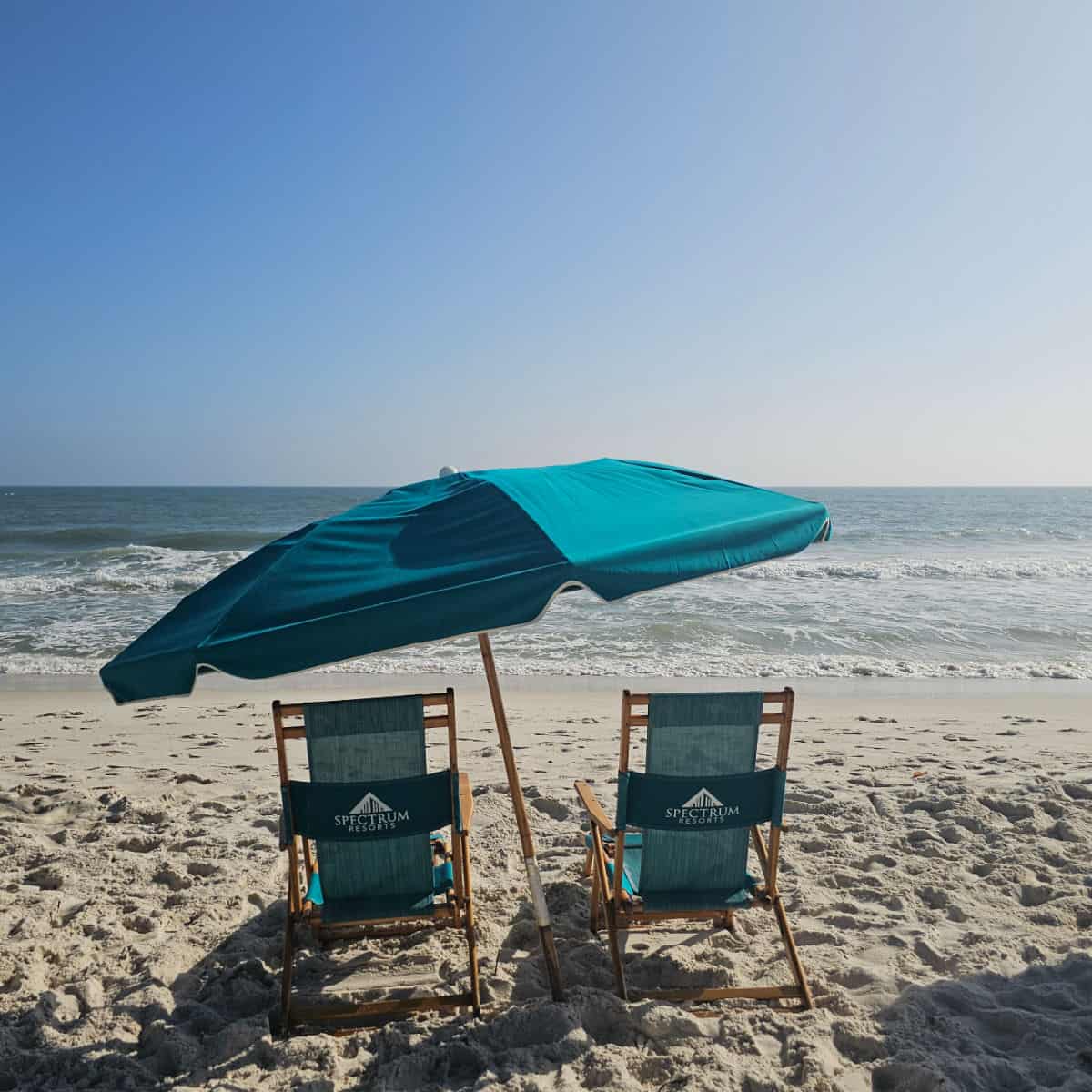 Two turquoise Spectrum chairs and a turquoise umbrella on the beach with calm Gulf waters behind them.