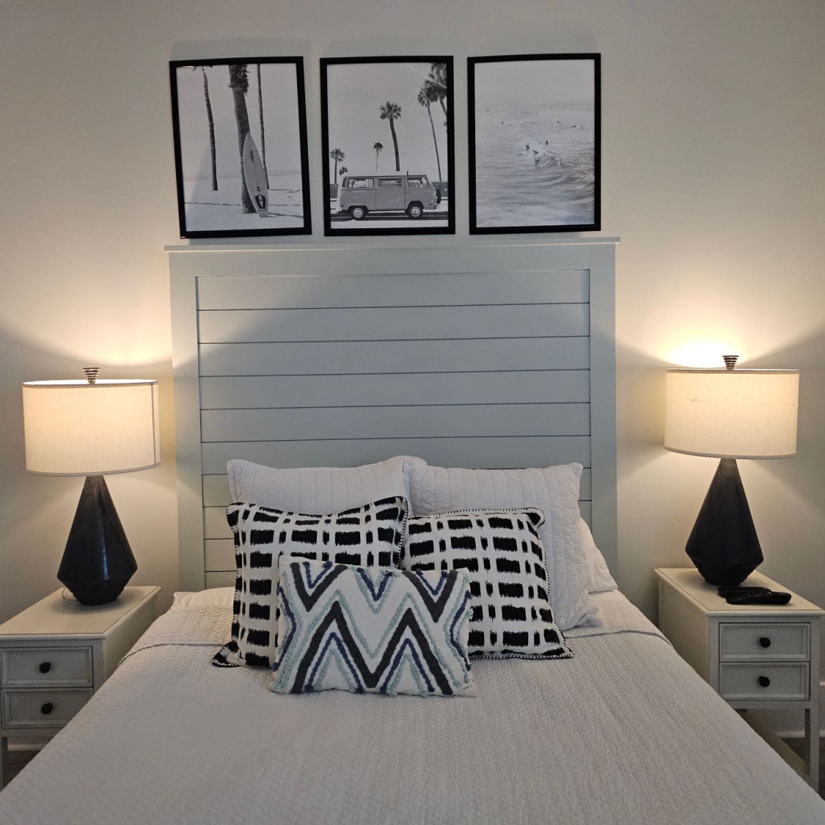 Bed with wooden headboard, white linens, black and white pillows, Black and white photos above the bed, white nightstand with two black lampbases and white shades