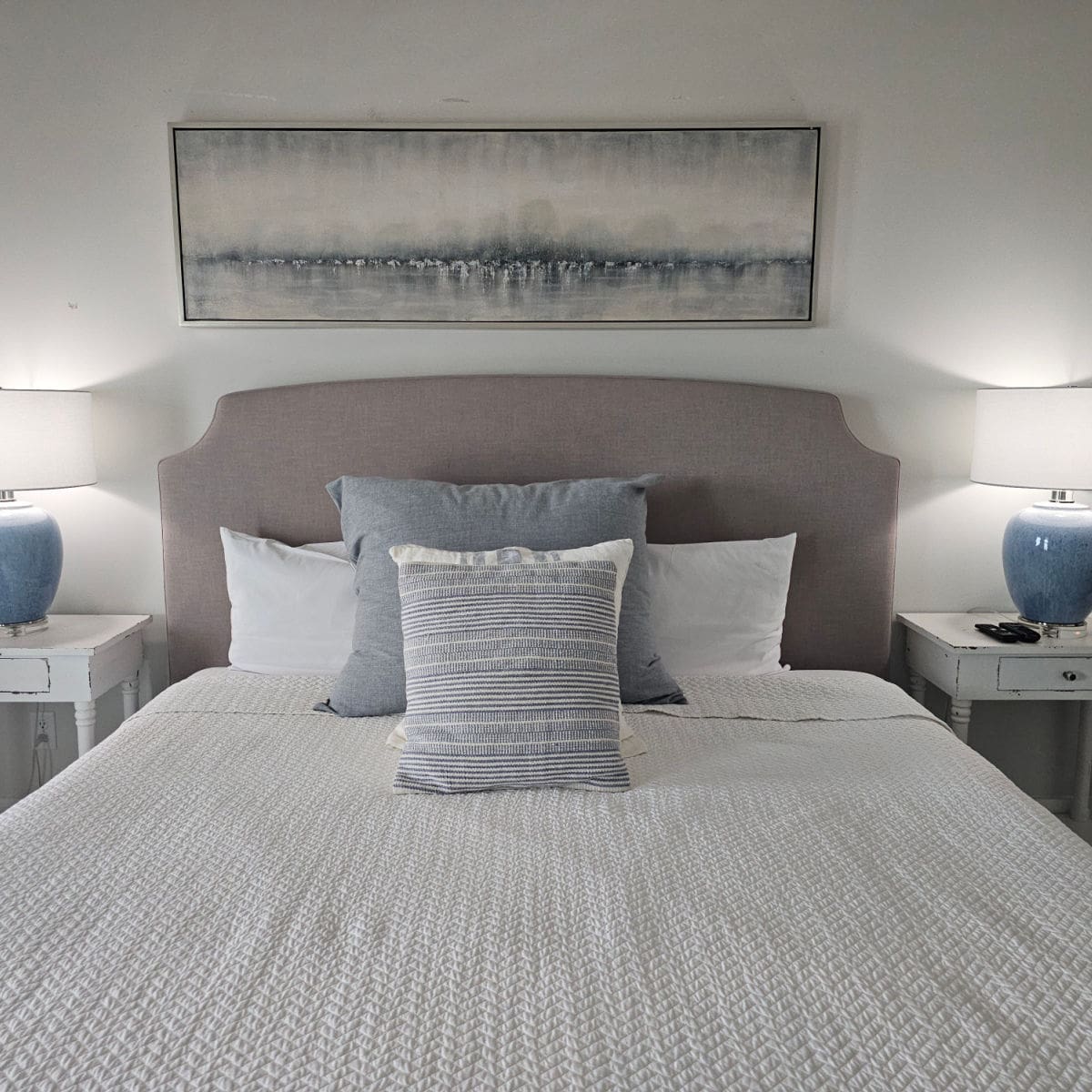 Bed with a brown headboard, white linens, and grey pillows, two white nightstands with lamps and art above the bed