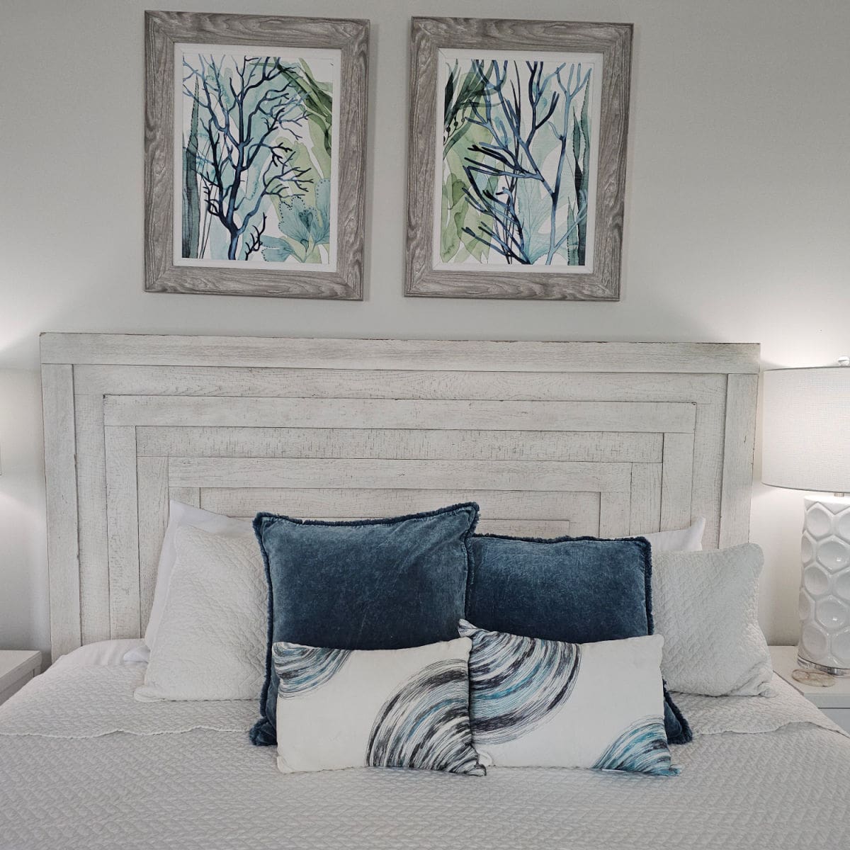 Bed with white wooden headboard, white linens, blue and white pillows. Art above the bed, white lamp and nightstand on the side