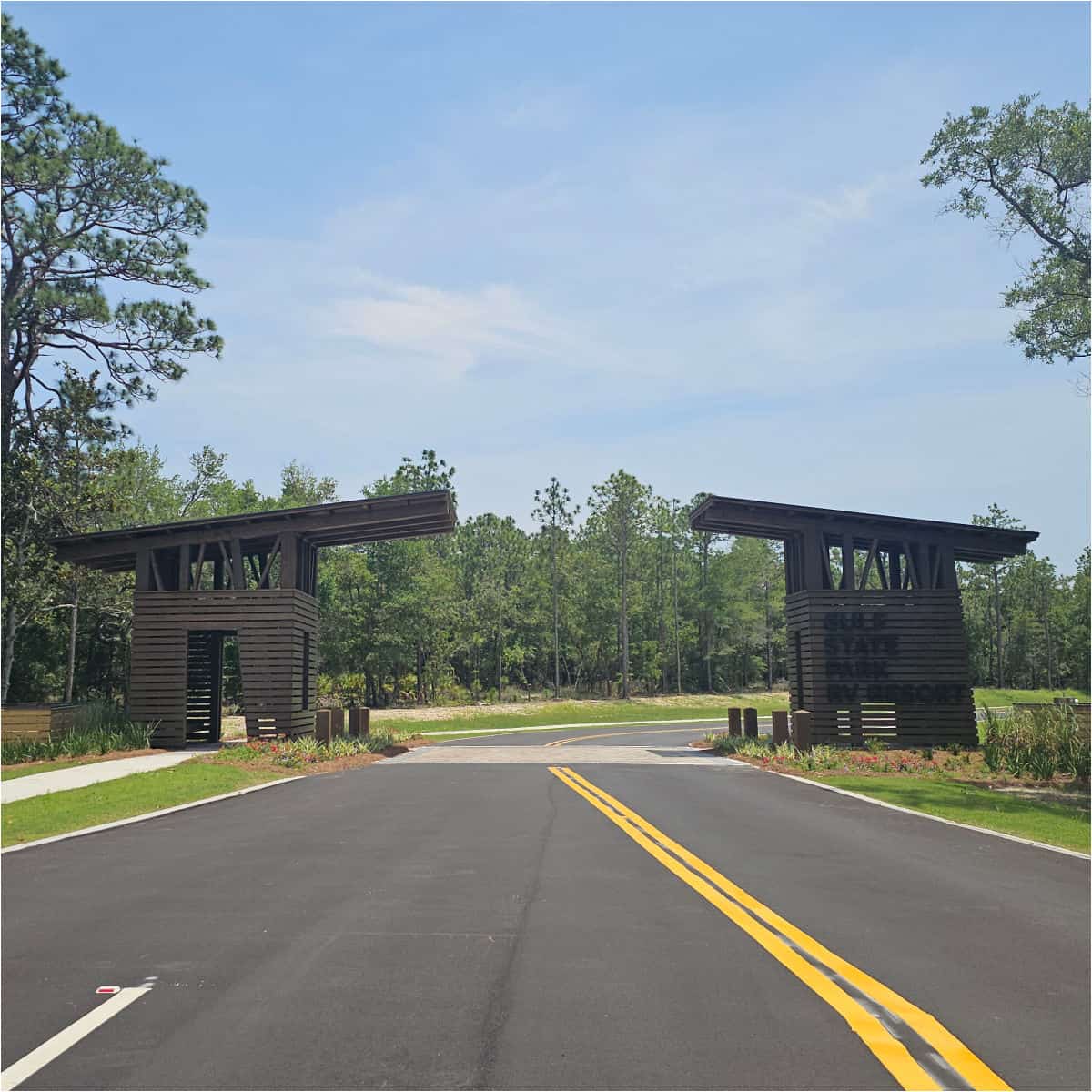 Entrance to Gulf State Park RV Resort with brown wooden structures on either side of the road, trees in the background