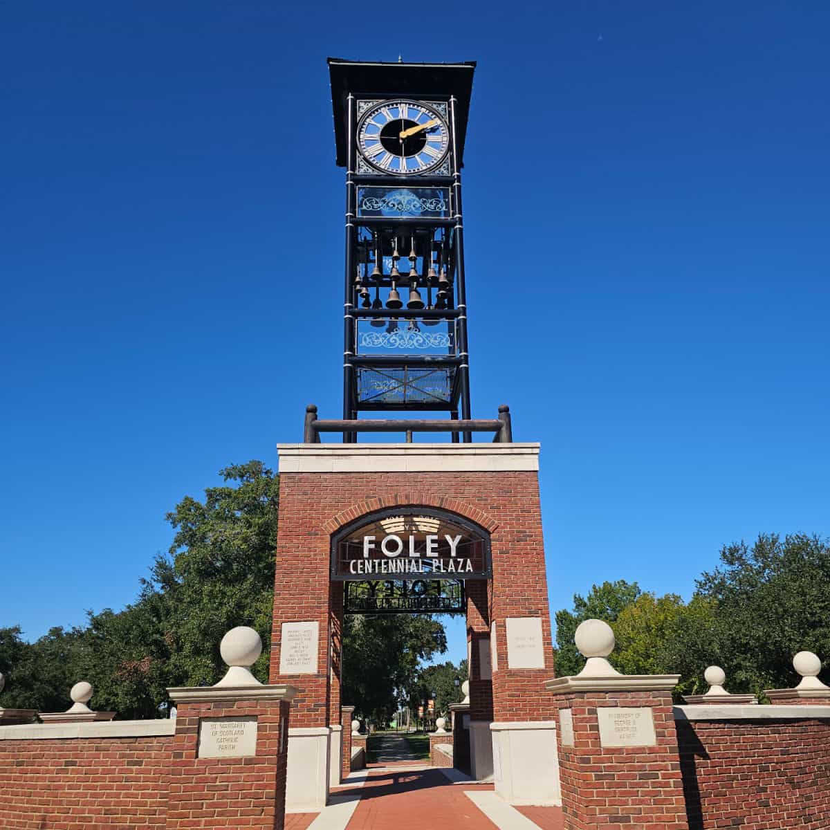 Clock tower with Foley Centennial Plaza sign at the base, surrounded by a brick wall