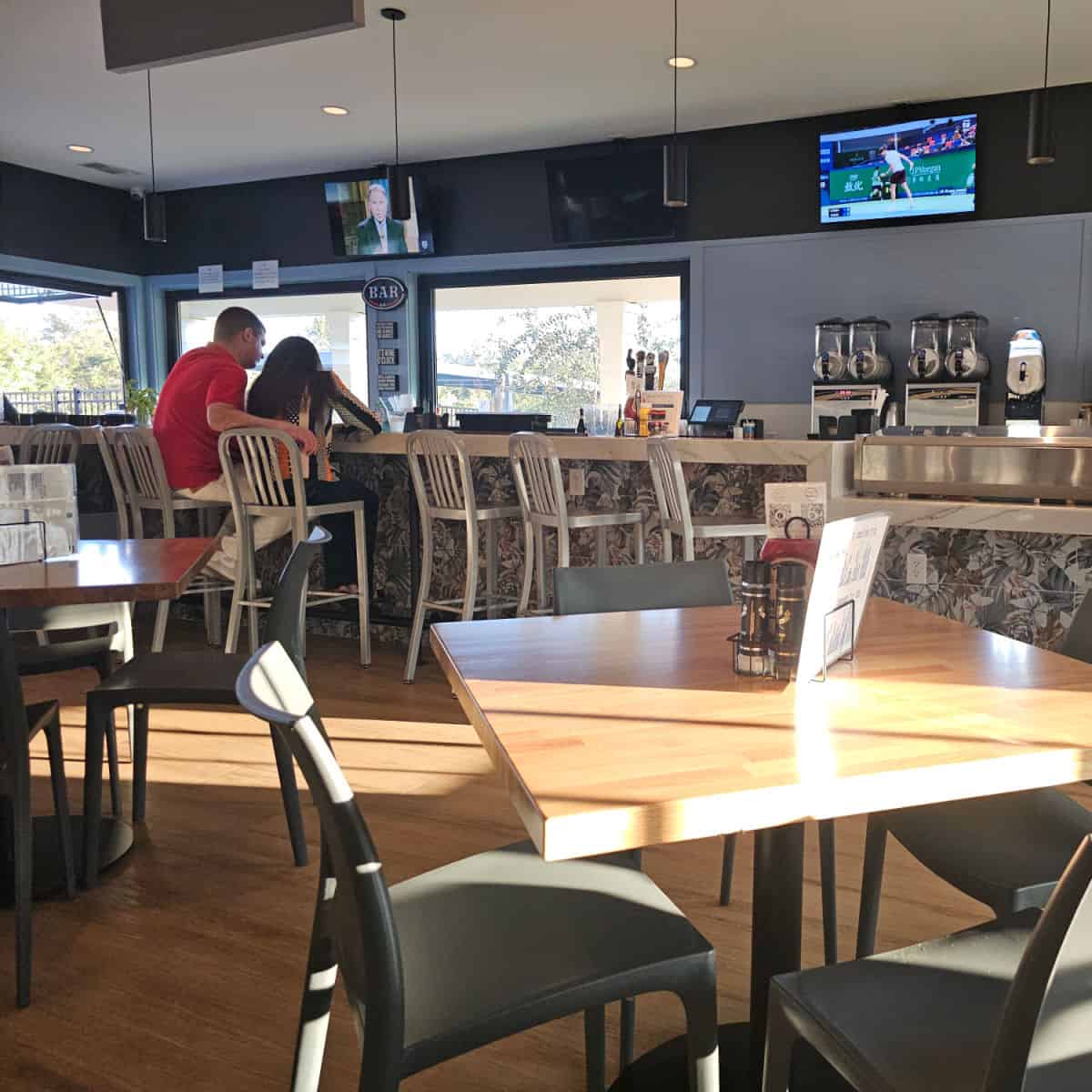 indoor seating with tables and chairs, long bar with silver bar chairs, flatscreen tvs above the bar and drink mixers along the wall
