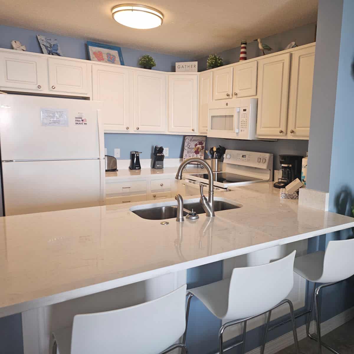 Looking into the kitchen with white cabinets and white appliances, coastal art on top of the cabinets. white stools under the counter