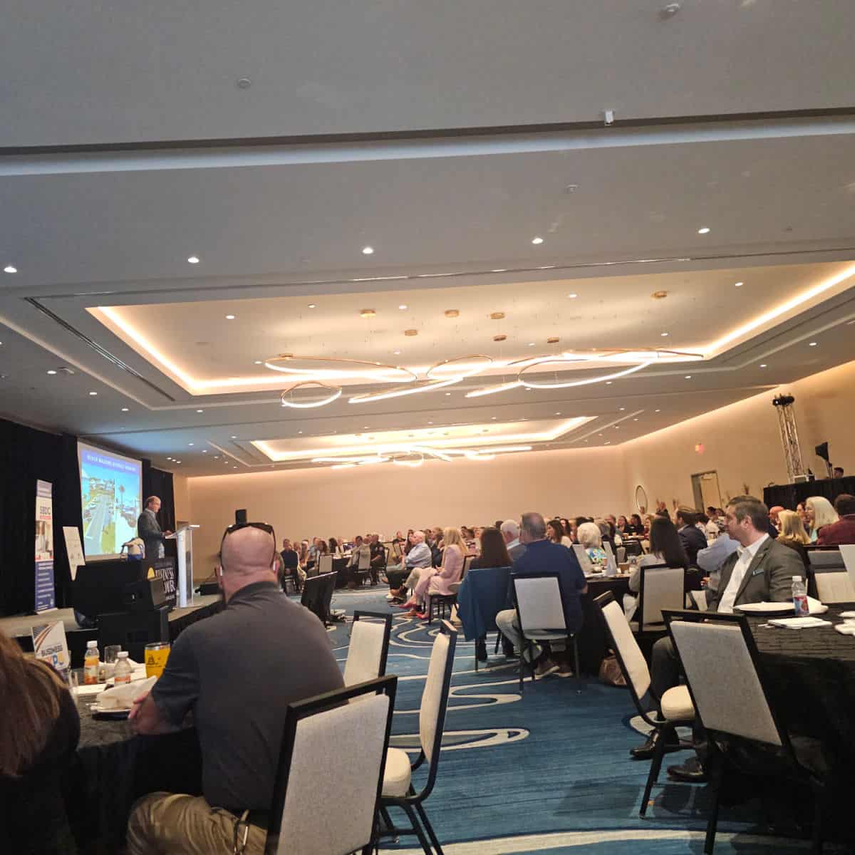 Large meeting room with people at round tables, speaker at the front with a video screen behind them.