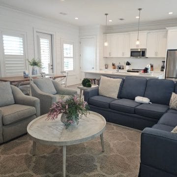 Living room with a blue couch and two grey chairs, circle table in the middle with a floral arrangement, white kitchen in the background with stainless steel appliances