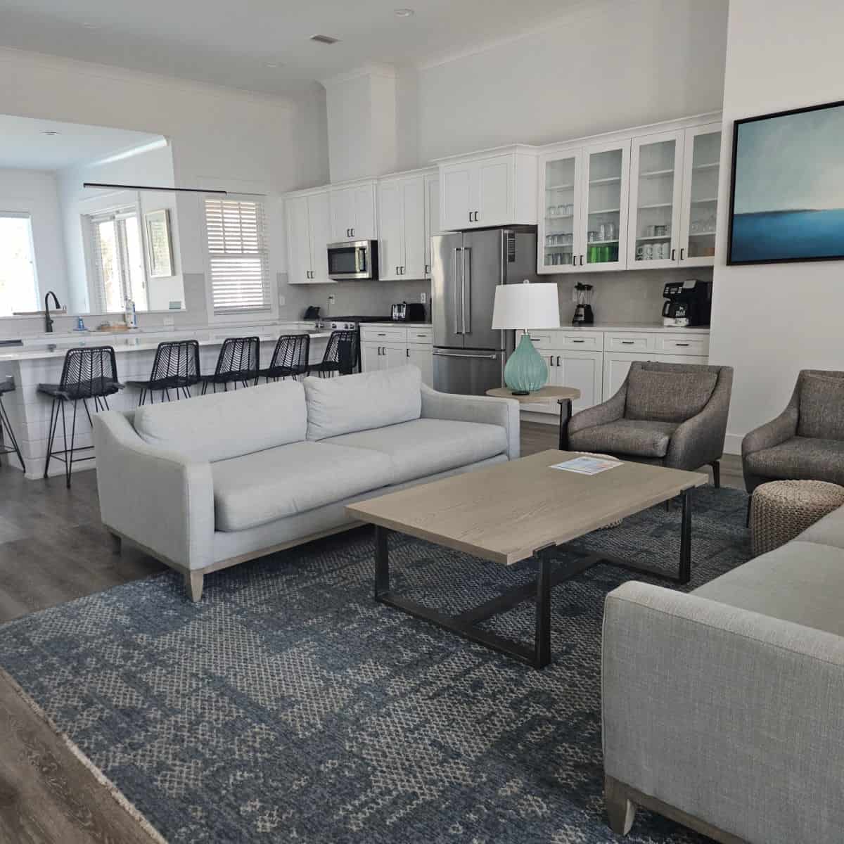 Living room and kitchen area with two grey couches, table in the middle of the couches, two chairs on the side, looking towards a white kitchen with stainless steel appliances, dark stools against the white counter island.