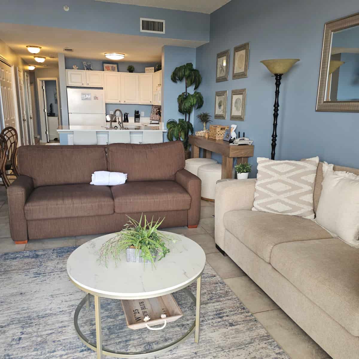 Looking from the living room with a brown couch and cream colored couch, circle table in the middle, towards the kitchen with white cabinets and white appliances, blue walls in both rooms