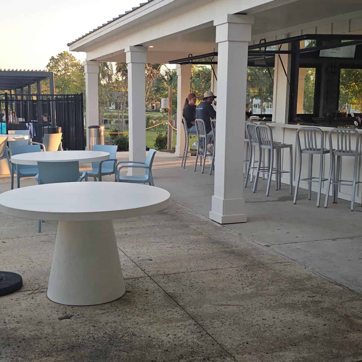 outdoor seating with white tables and blue plastic chairs in the open, under the cover of the restaurant, there are tall silver bar stools next to an open bar window