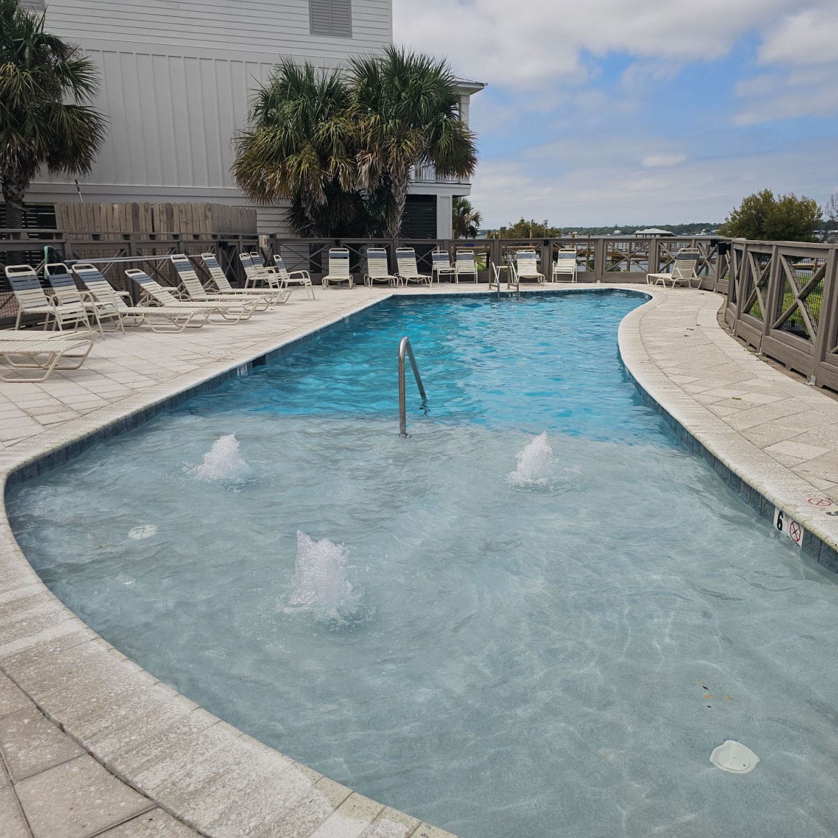 Outdoor pool with bubbles in the shallow end, lounge chairs spread around the pool deck, fence around the whole area.